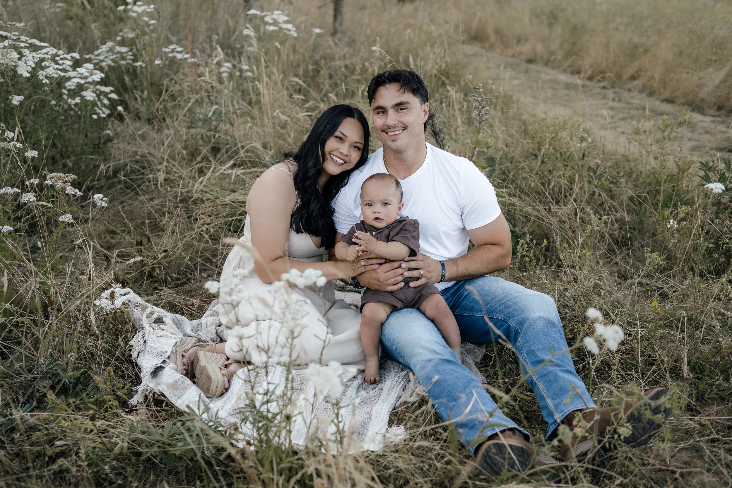 A happy family of three sitting on a blanket in a field of tall grass and wildflowers, smiling at the camera.