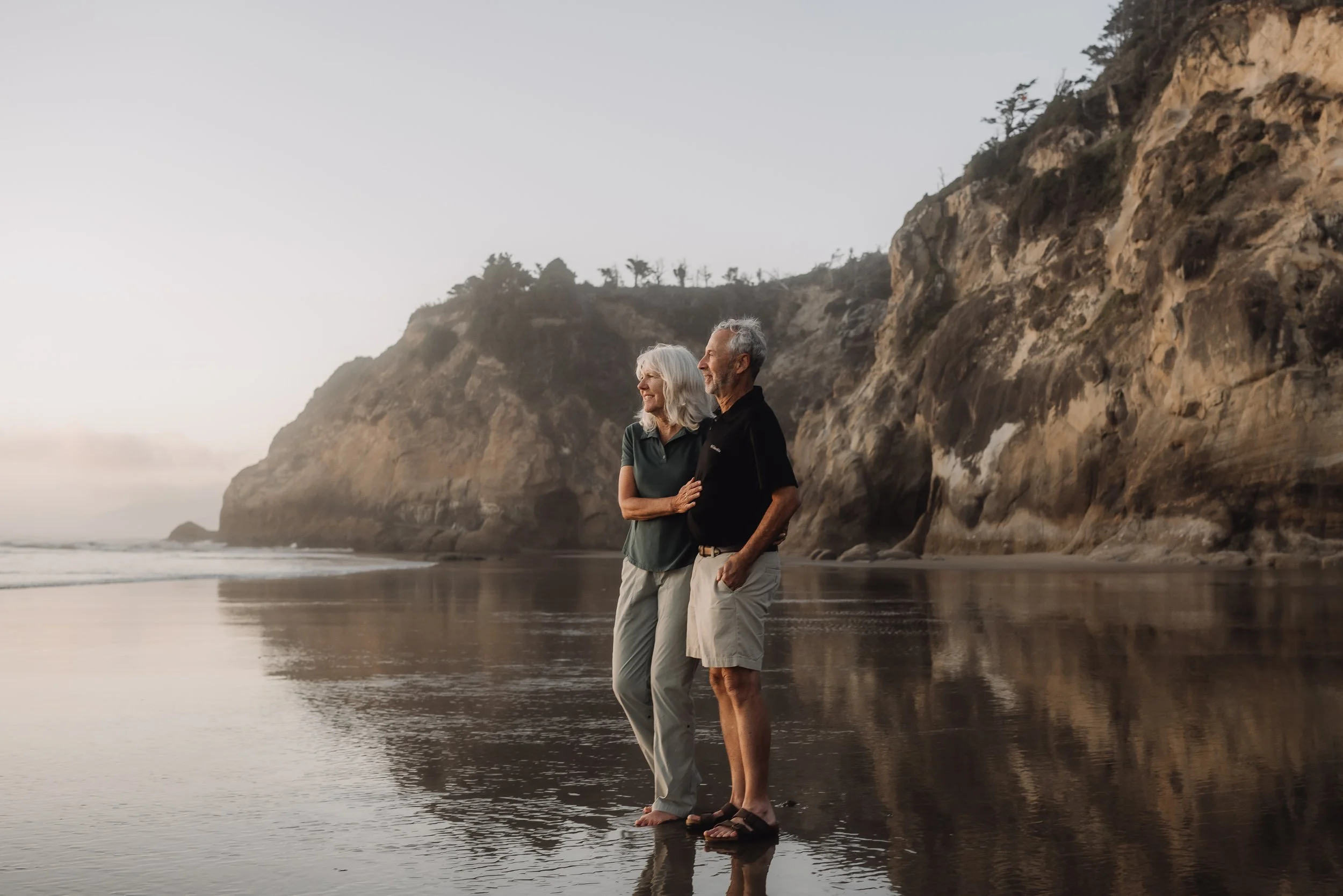 An elderly couple walking on the beach at sunset with cliffs and ocean in the background.