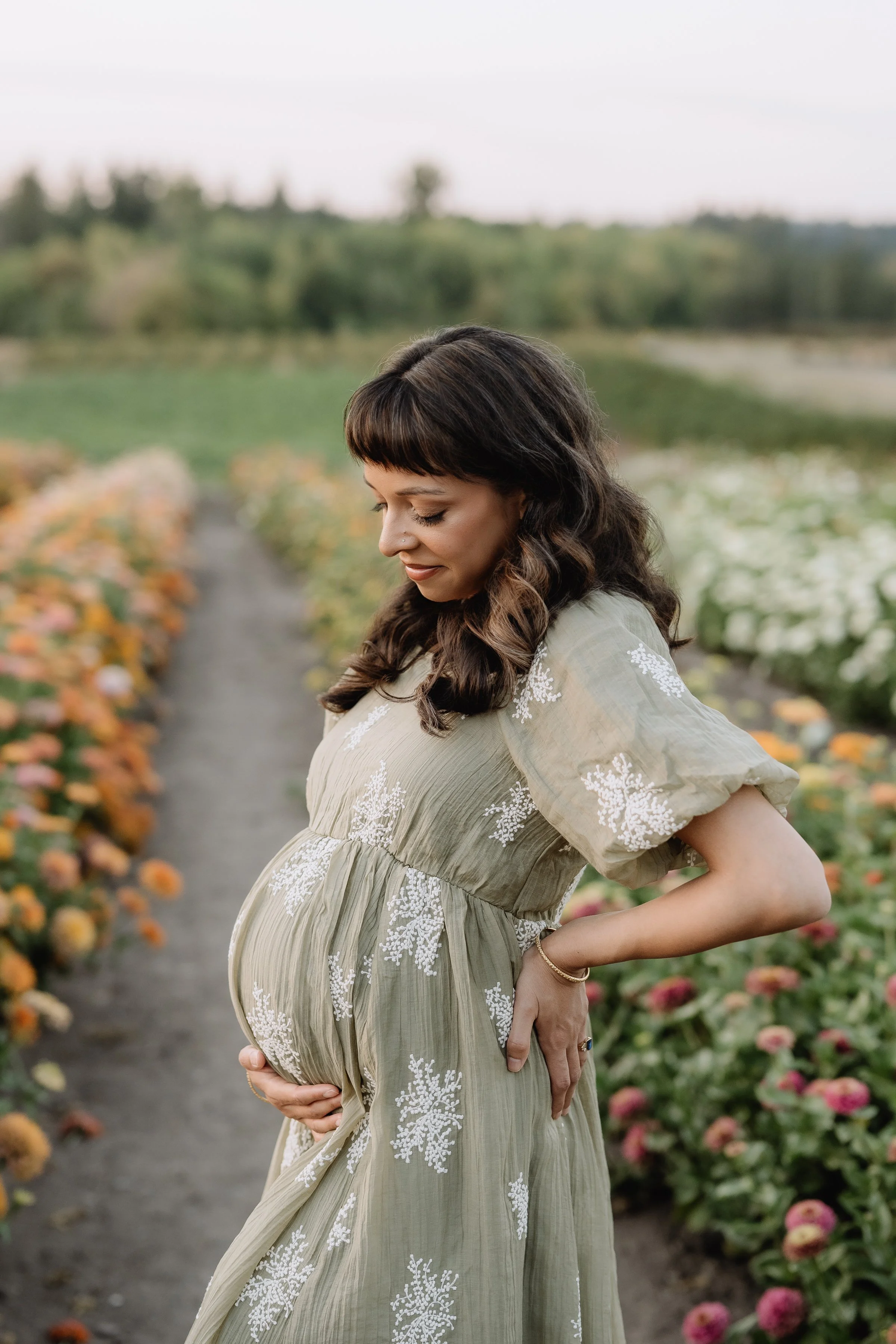 Pregnant woman in a flowy dress standing in a flower field, gently cradling her belly, with greenery and flowers in the background.