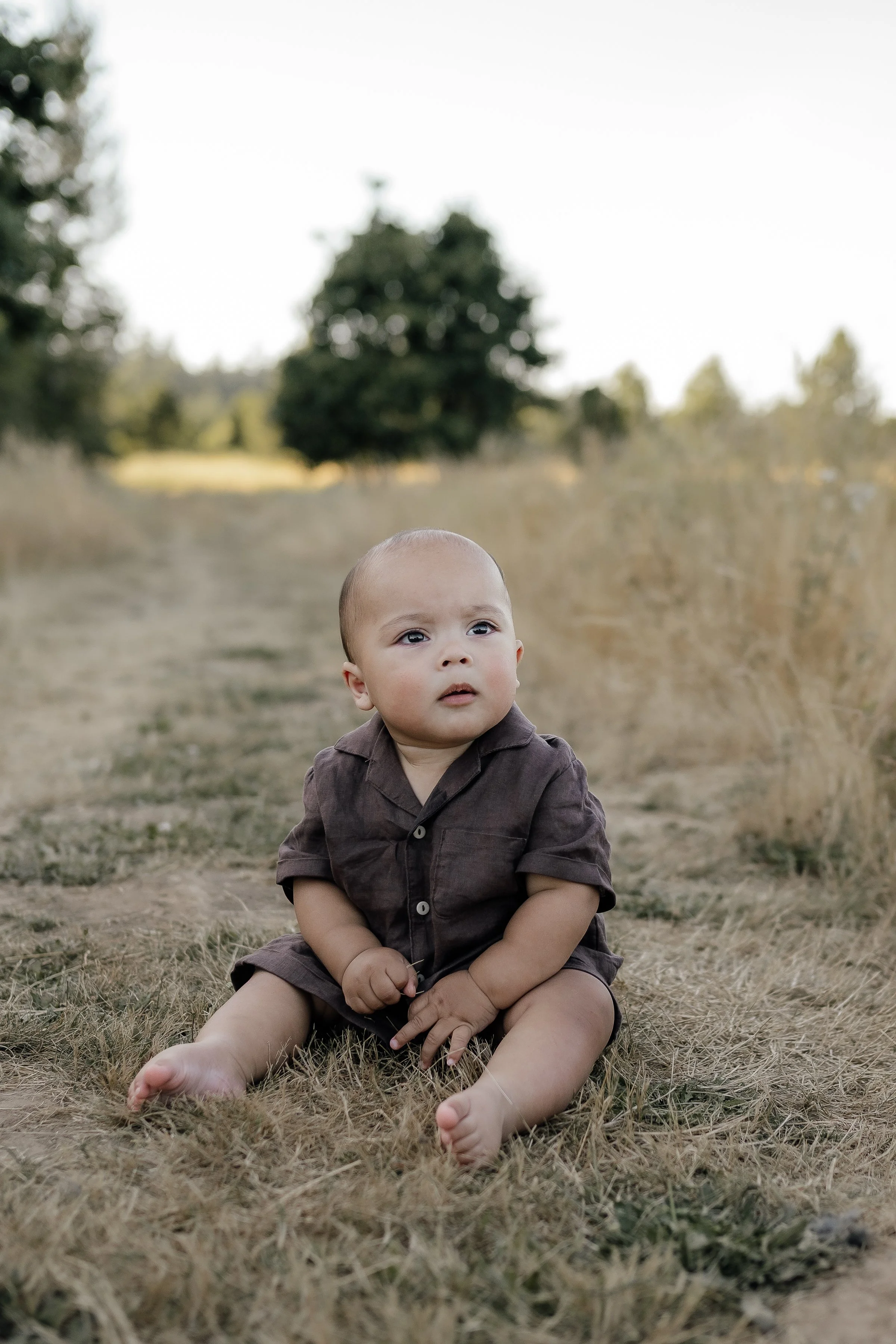 Young baby sitting outdoors on a dry grassy path with trees in the background.