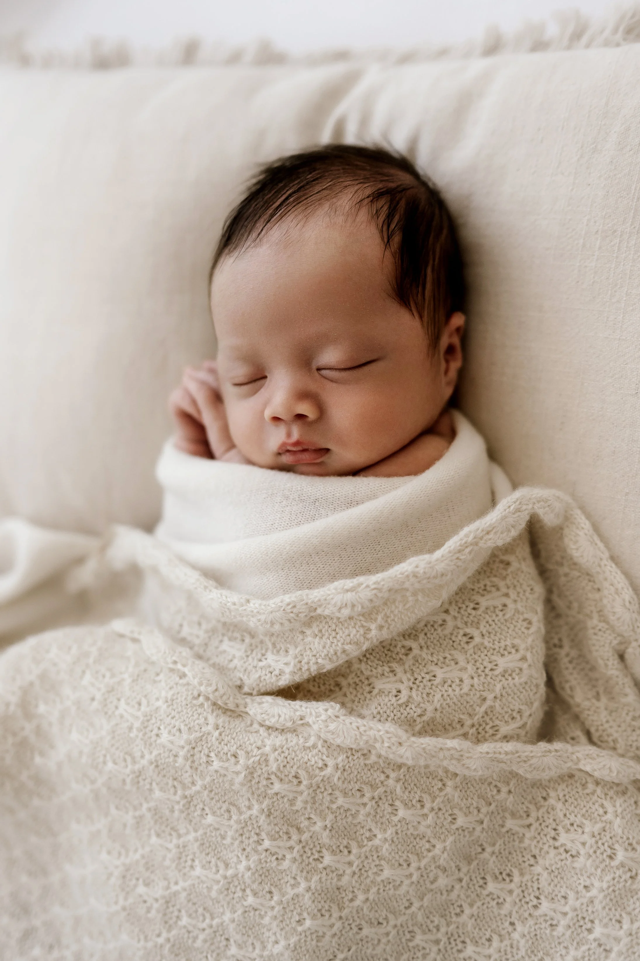 A sleeping newborn baby wrapped in a cream-colored, knitted blanket, resting against a light-colored couch.