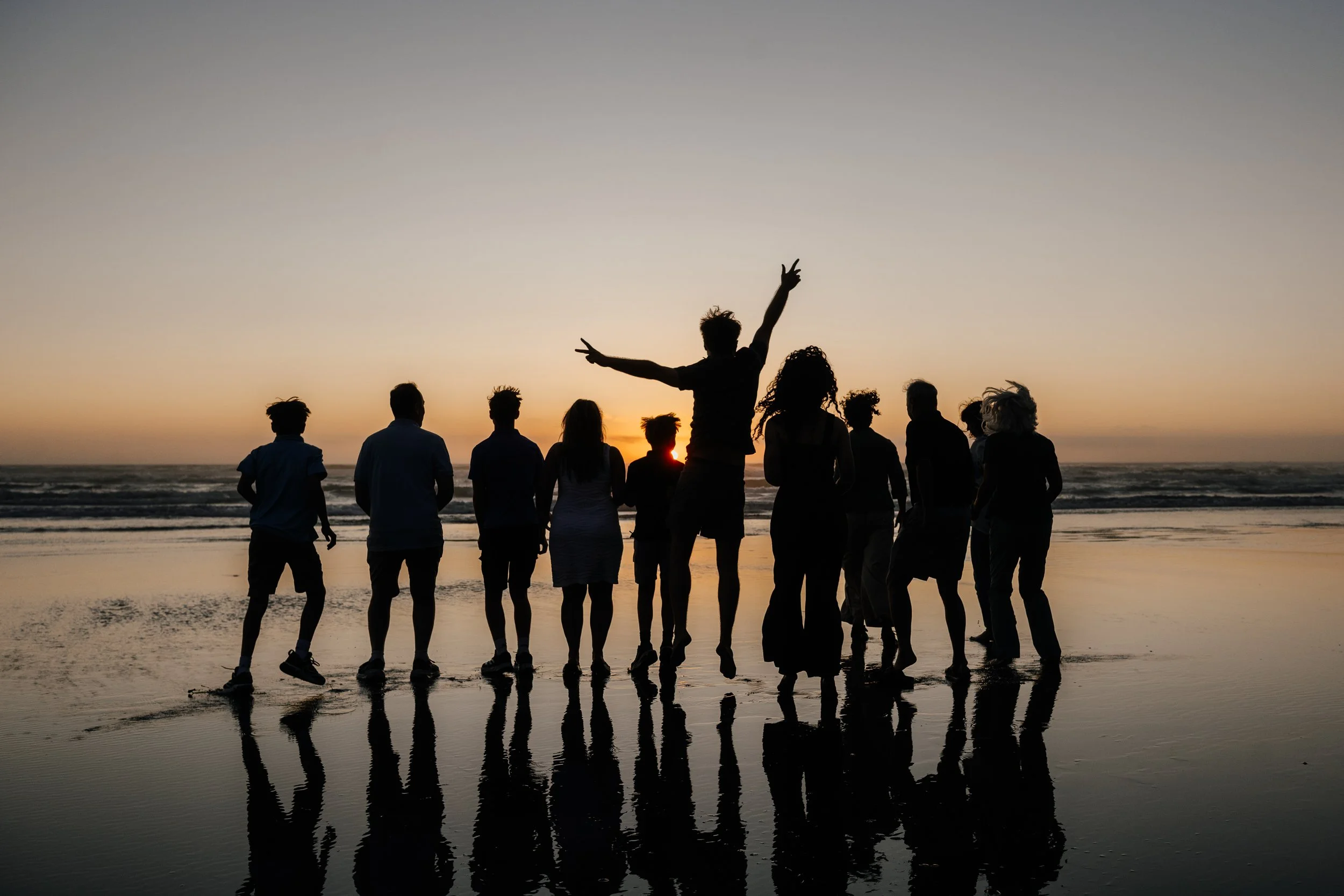 Group of people standing on the beach at sunset, some jumping and some standing still, with their reflections visible on the wet sand.