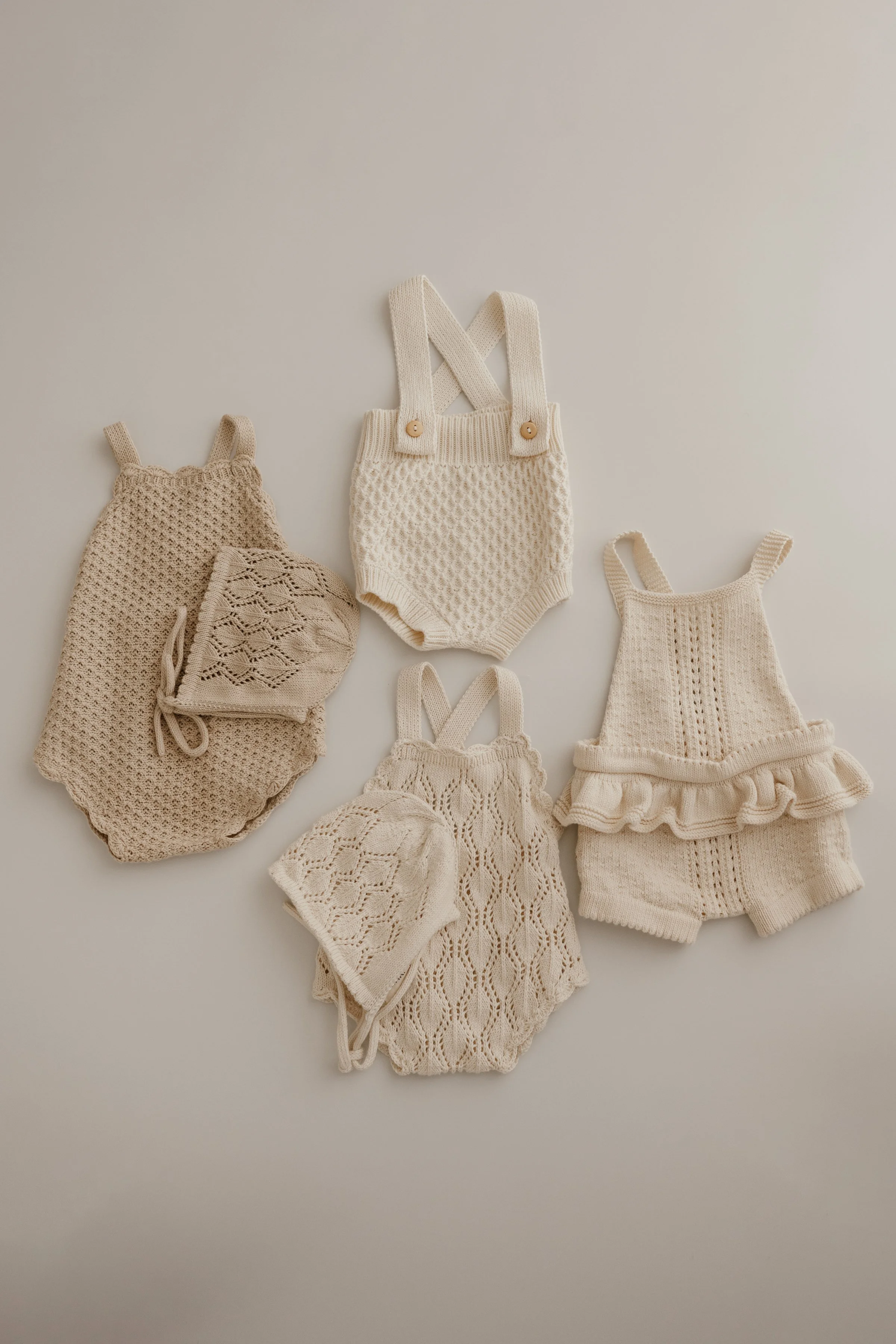 Collection of cream-colored knitted baby clothes, including sweaters, a pair of overalls, hats, and shorts, displayed on a plain background.