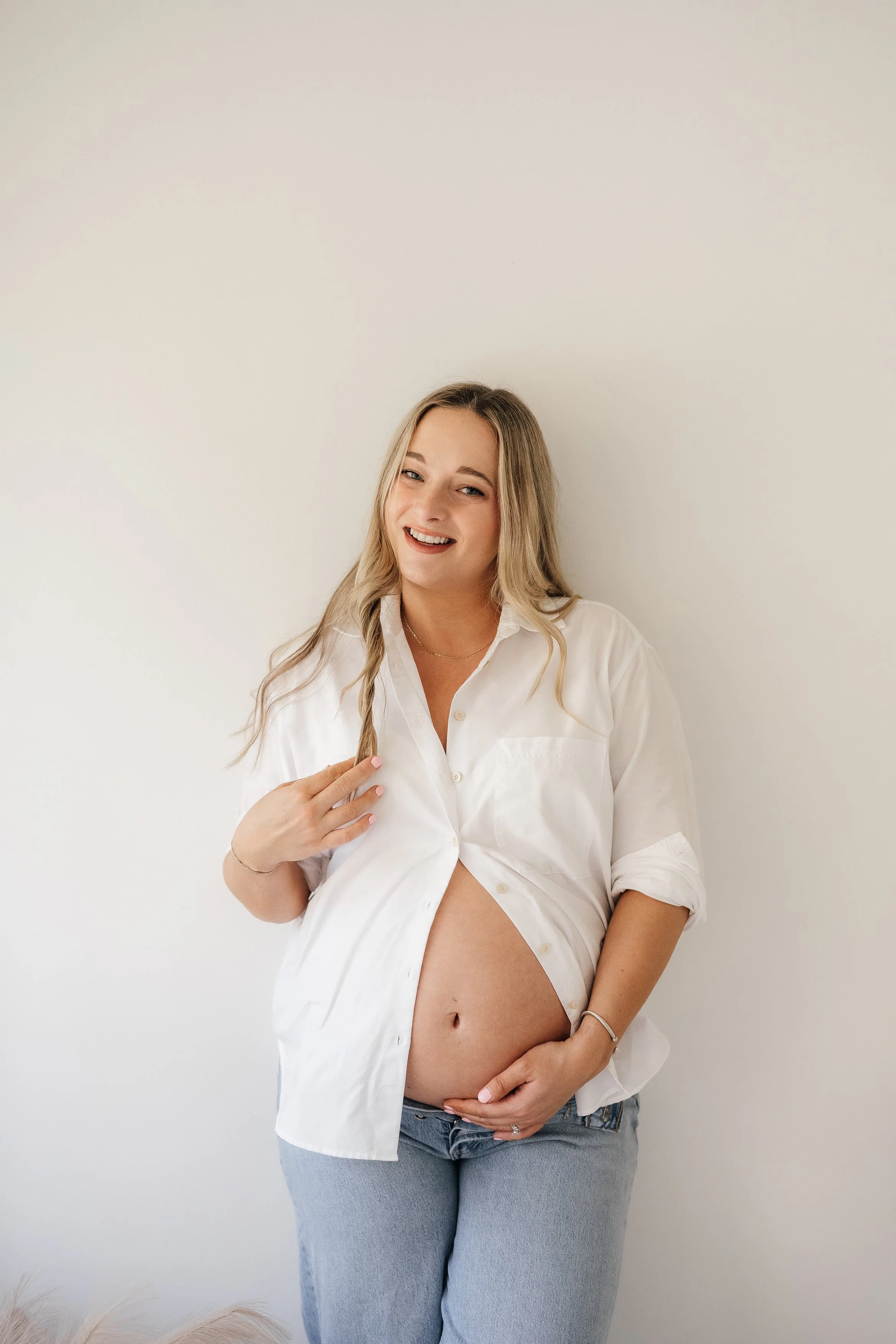 A pregnant woman with blonde hair smiling, wearing a white button-up shirt partially open, and light blue jeans, standing against a plain white wall.