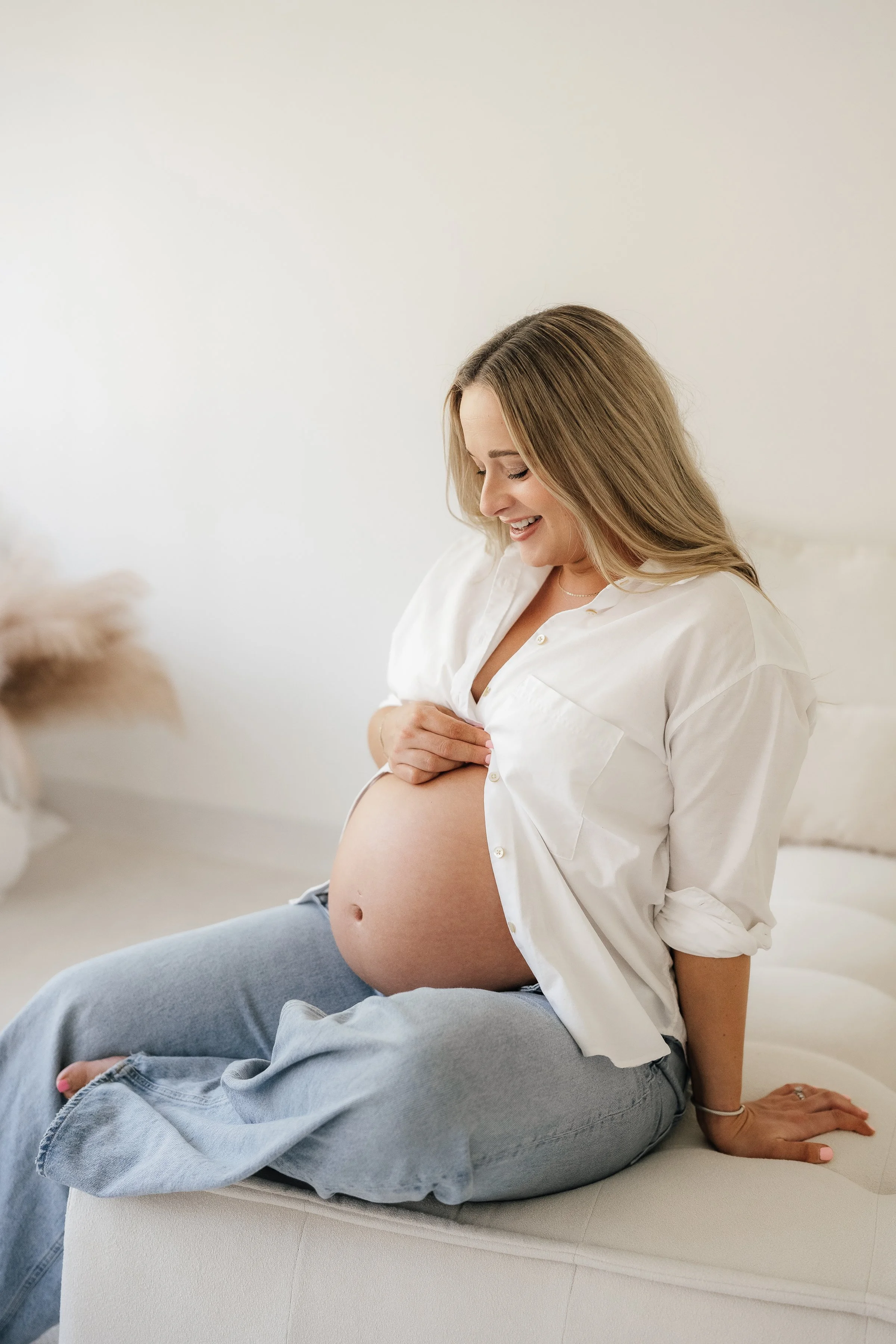 Pregnant woman in a white shirt smiling and touching her belly while sitting on a bed in a bright room.
