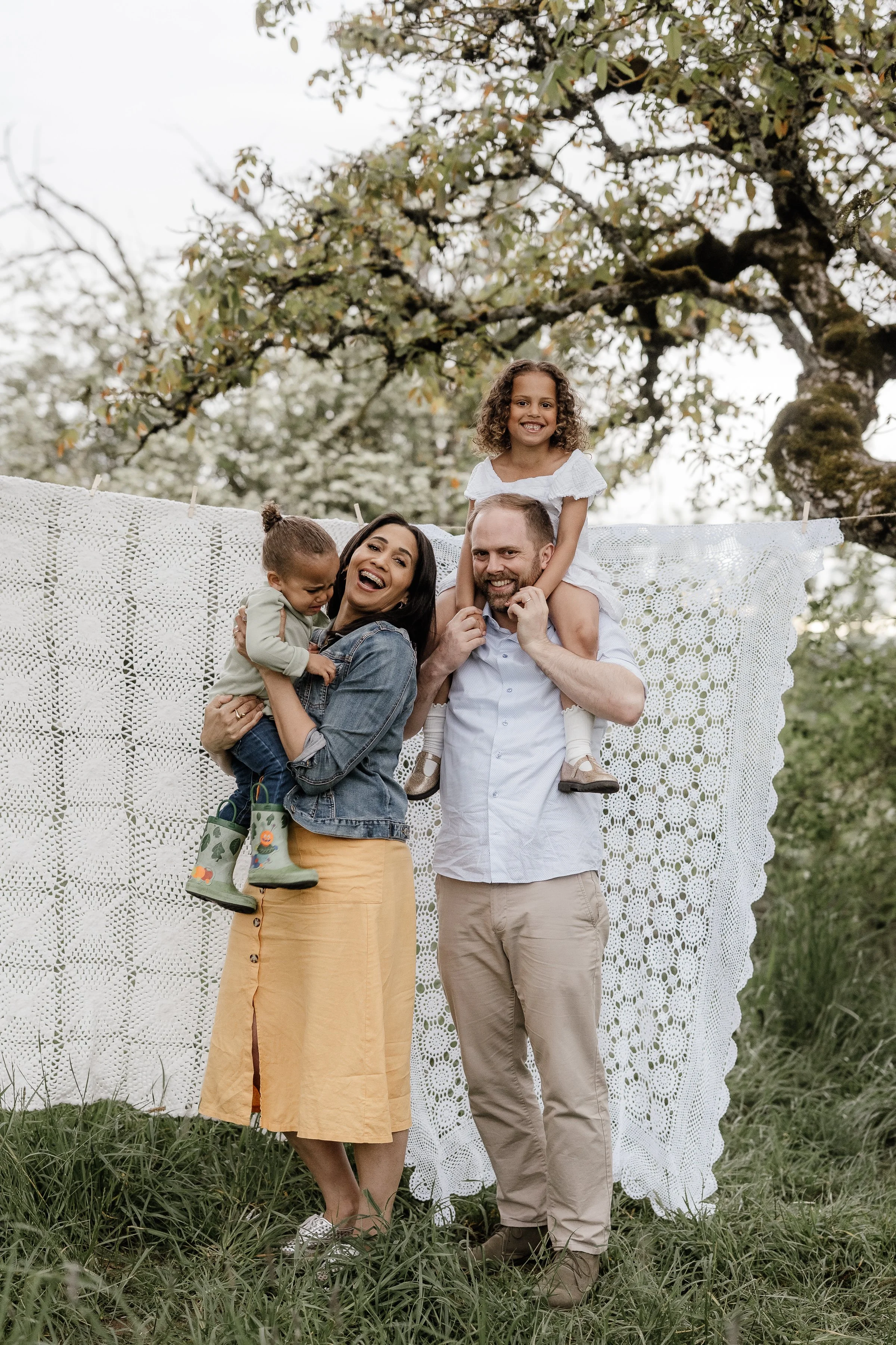 A family of four, including a woman, a man, and two young girls, smiling and playing outdoors on a grassy area with a large, crocheted white blanket hanging behind them and trees in the background.