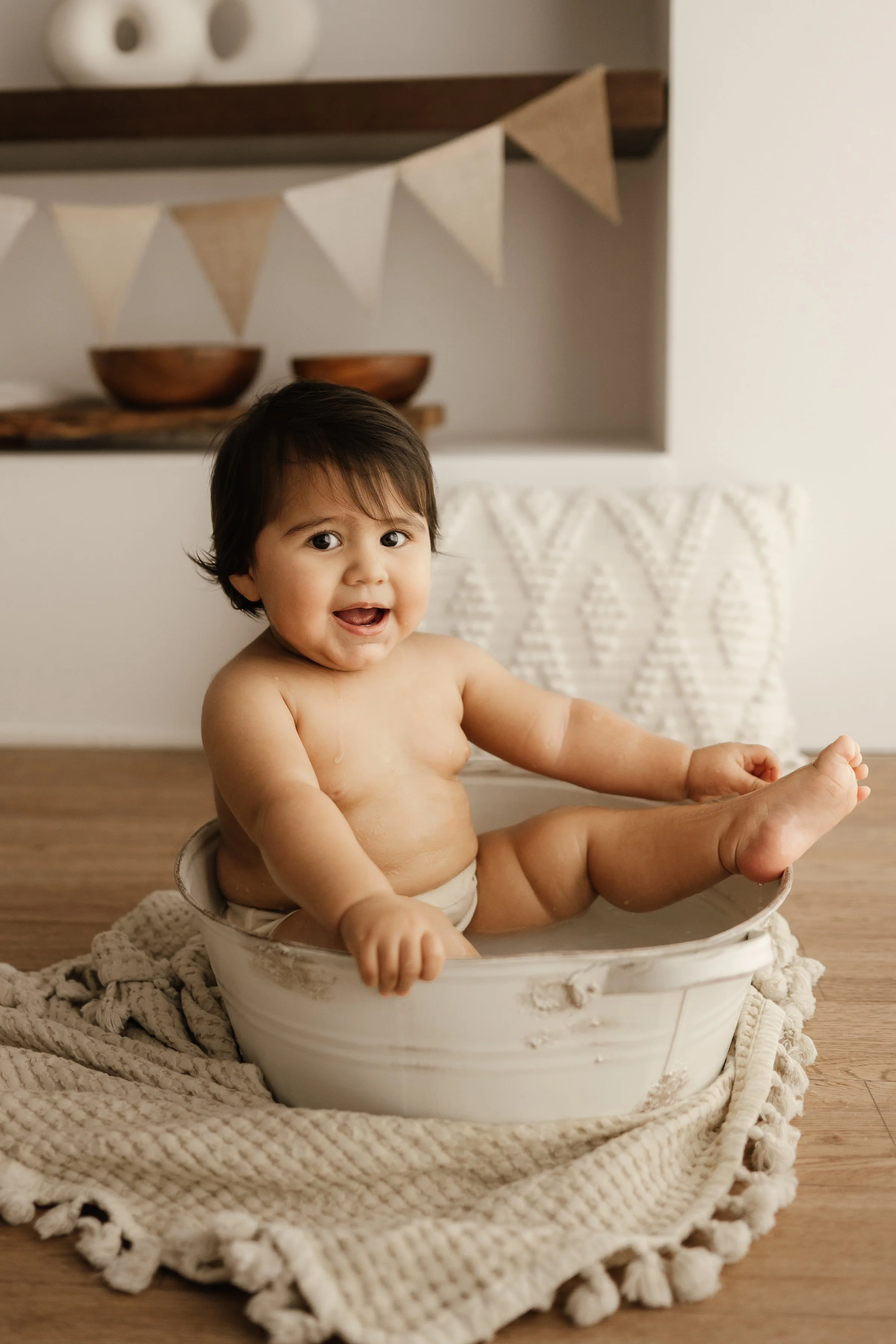 A smiling toddler sitting in a white ceramic basin, surrounded by a beige textured blanket, during bath time.