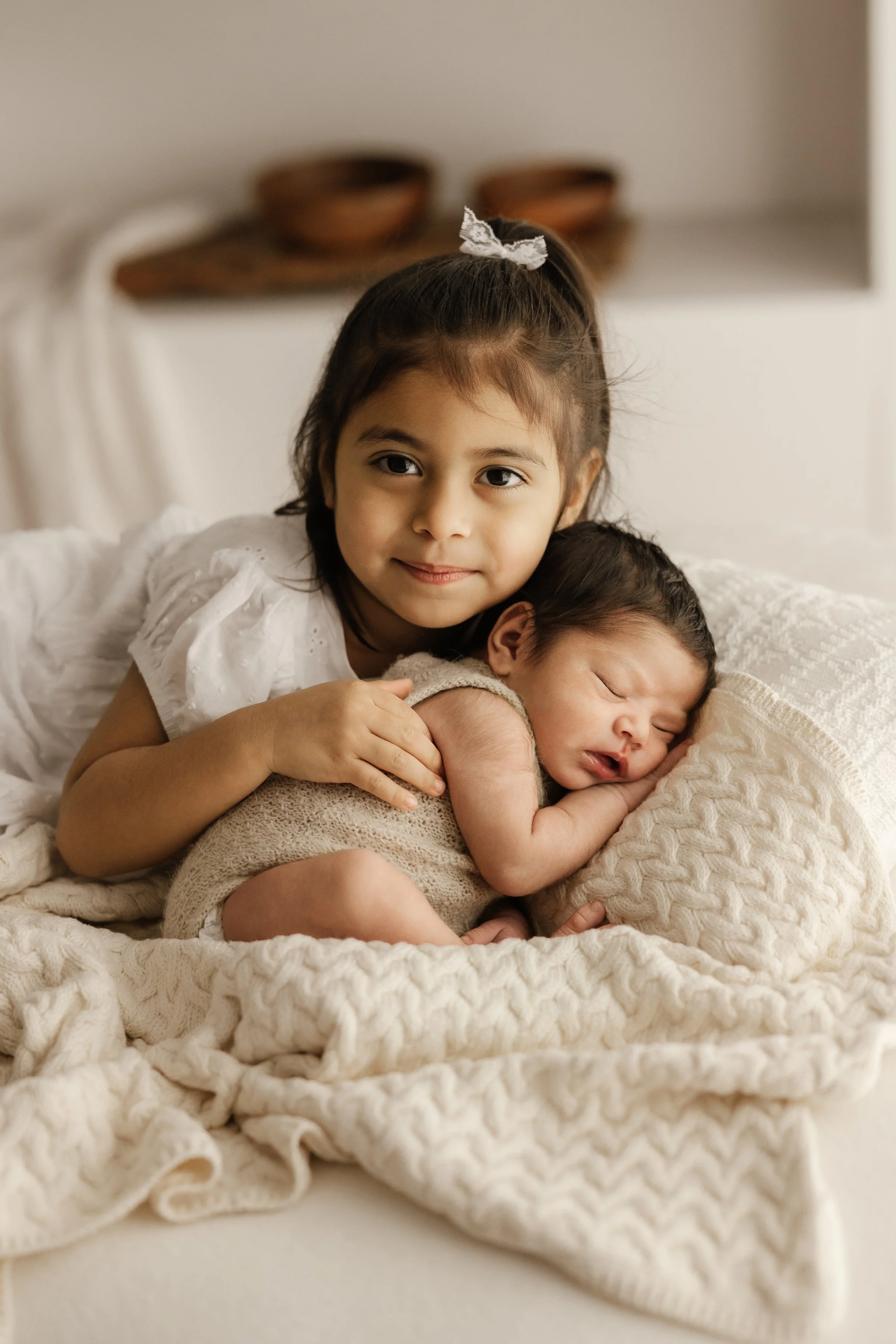 A young girl cuddling a sleeping toddler on a bed with cream-colored blankets.