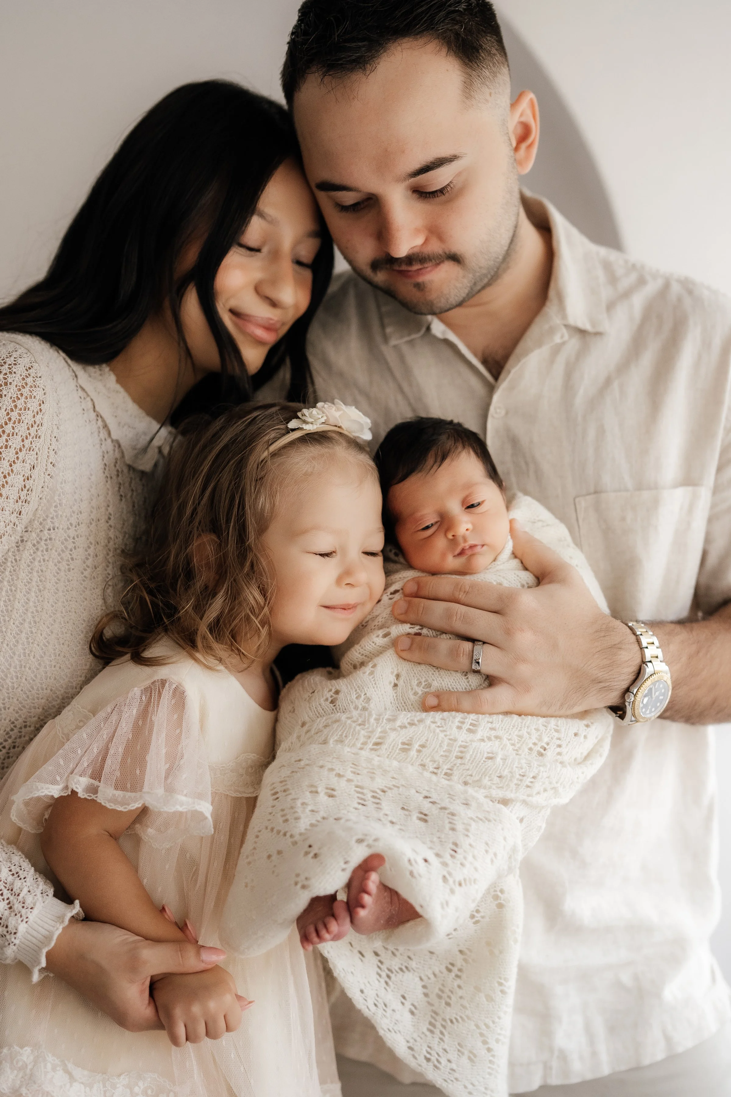 A family of four, including a woman, a man, a young girl, and a newborn, all dressed in light-colored clothing, gathered closely together, smiling and cuddling for a photo.