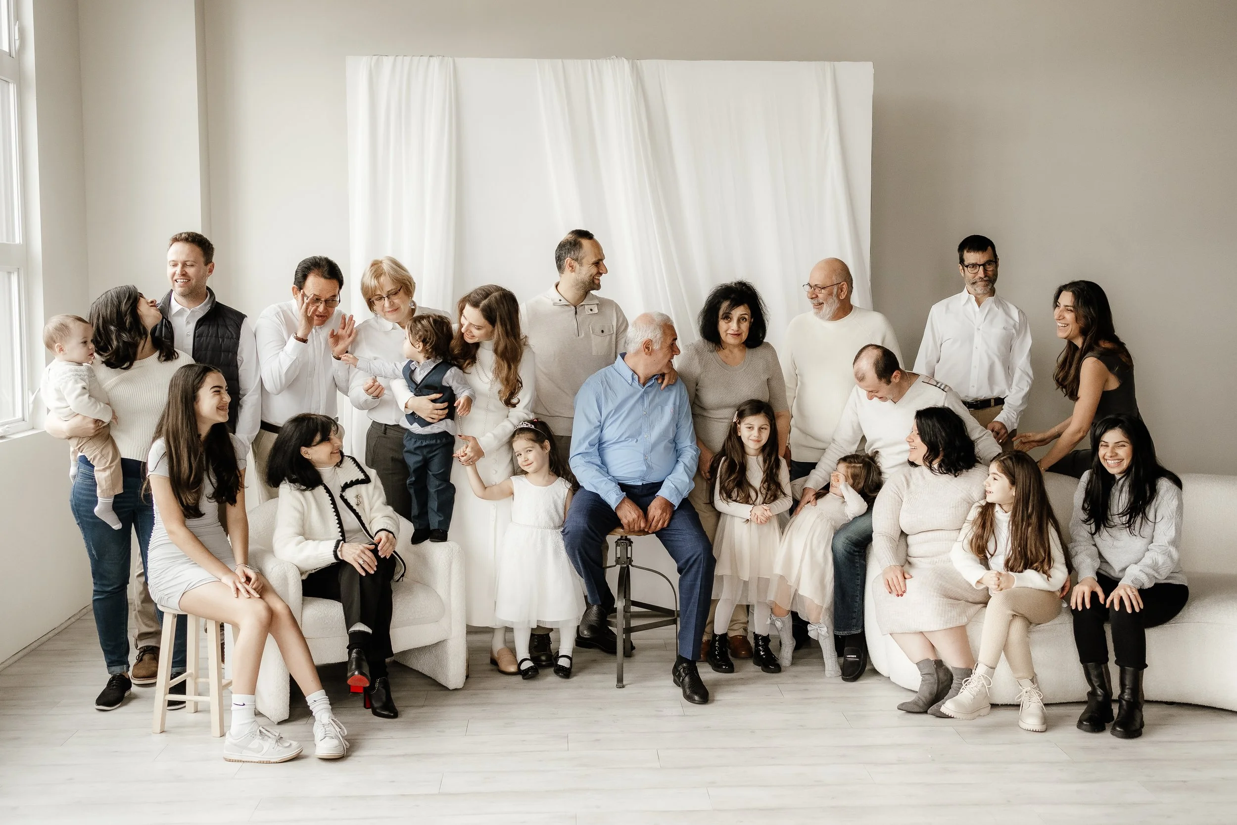 Large multi-generational family gathered in a bright living room, sitting and standing together, smiling and interacting with each other.
