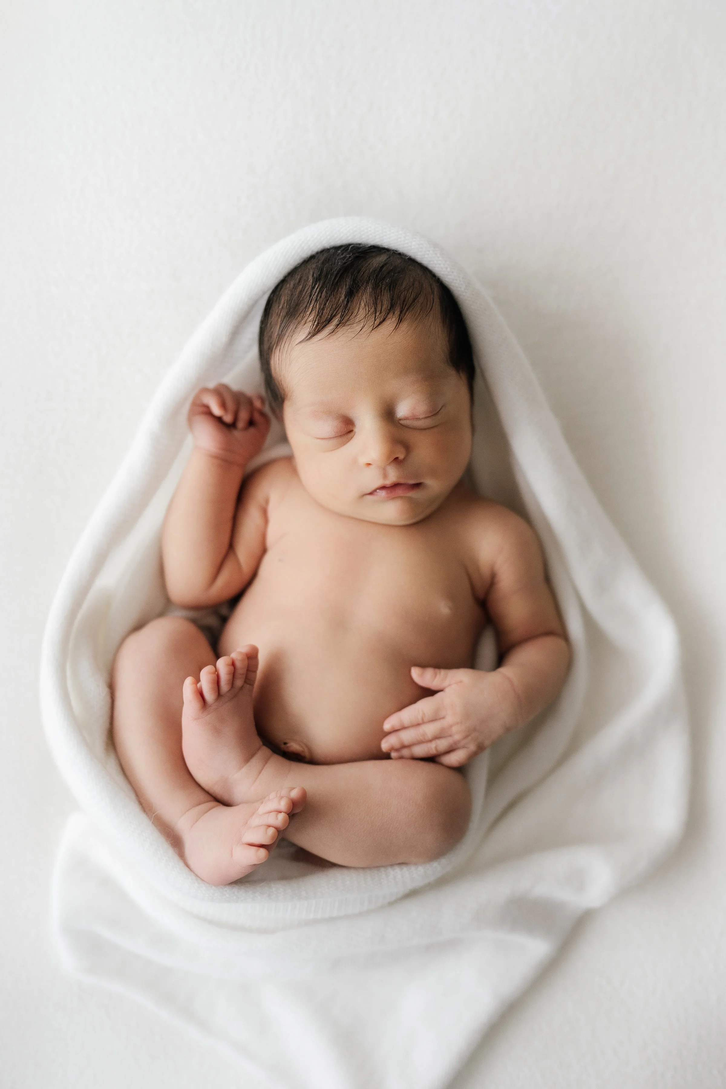 A sleeping newborn baby wrapped in a white blanket, lying on a white surface.