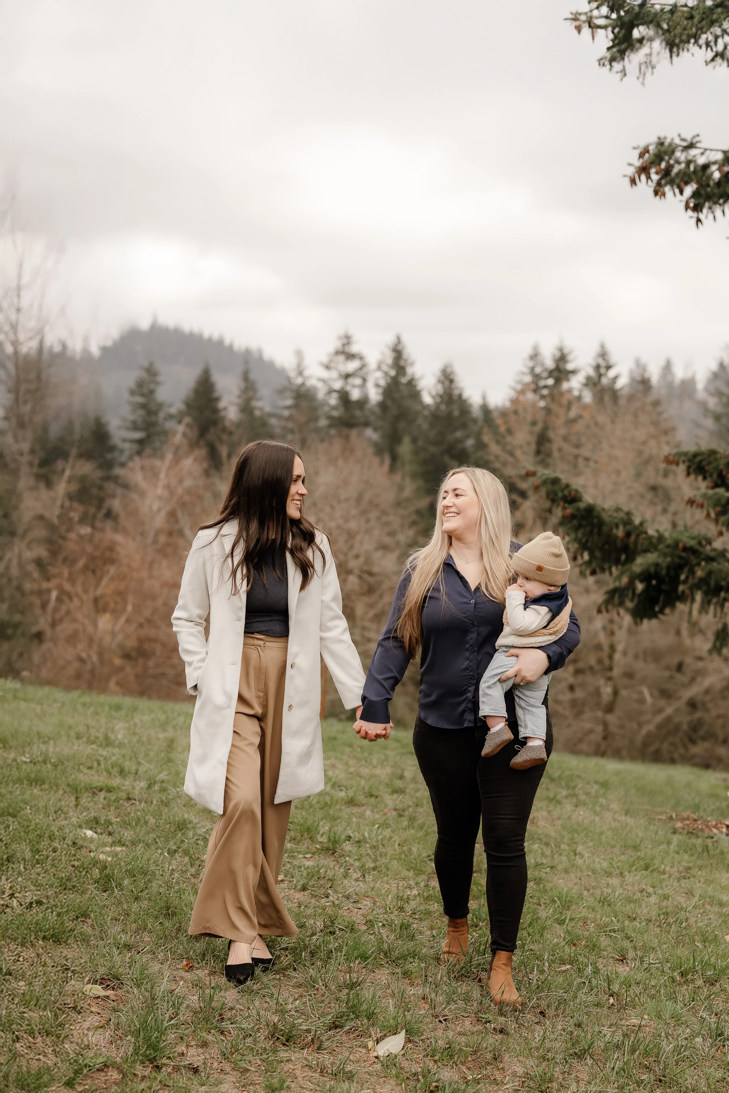 Two women walking outdoors in a park holding hands, smiling, with a child in one woman's arm, surrounded by trees and overcast sky.