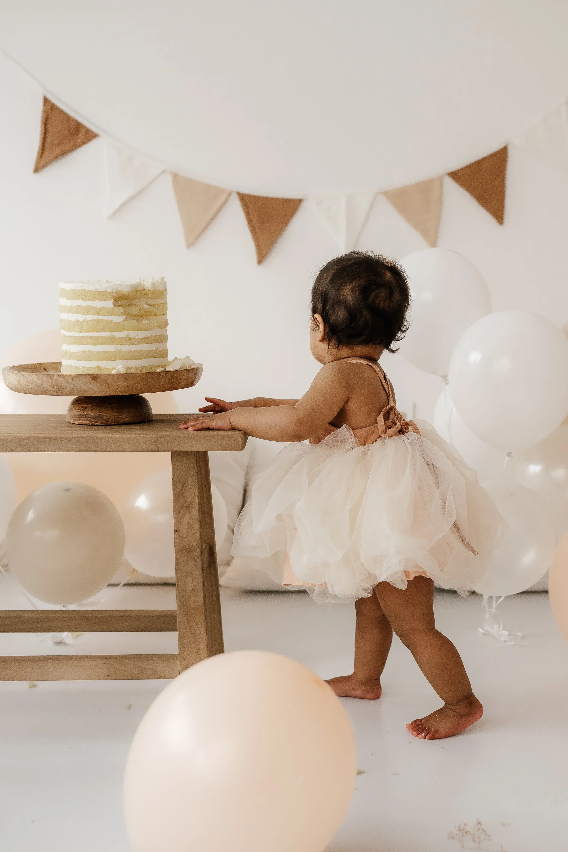 A baby girl in a pink tulle dress reaching toward a small cake on a wooden table, surrounded by beige and white balloons, with beige and white bunting hanging in the background.