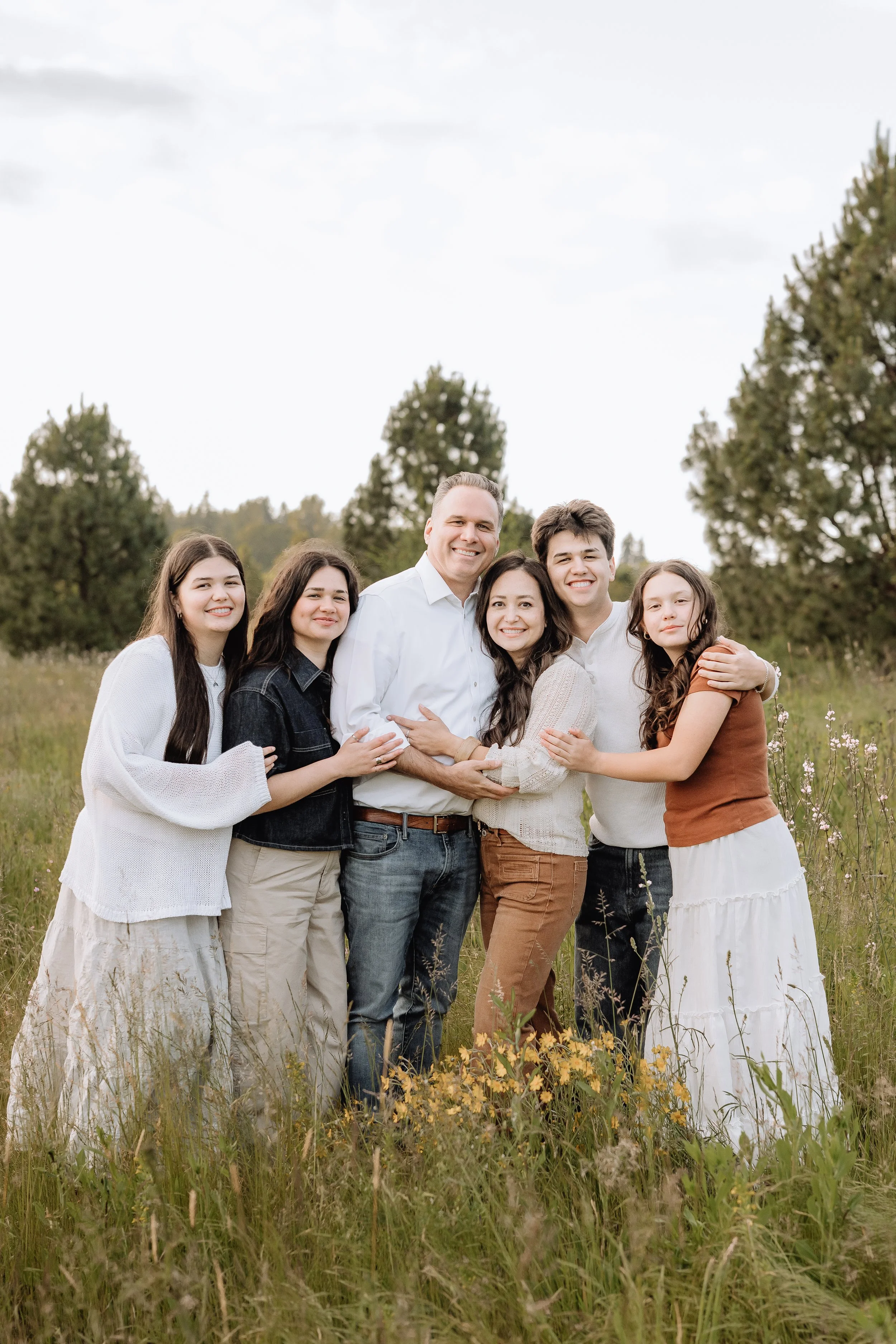 A multi-generational family of six people standing together outdoors in a grassy field with trees in the background, smiling and hugging.