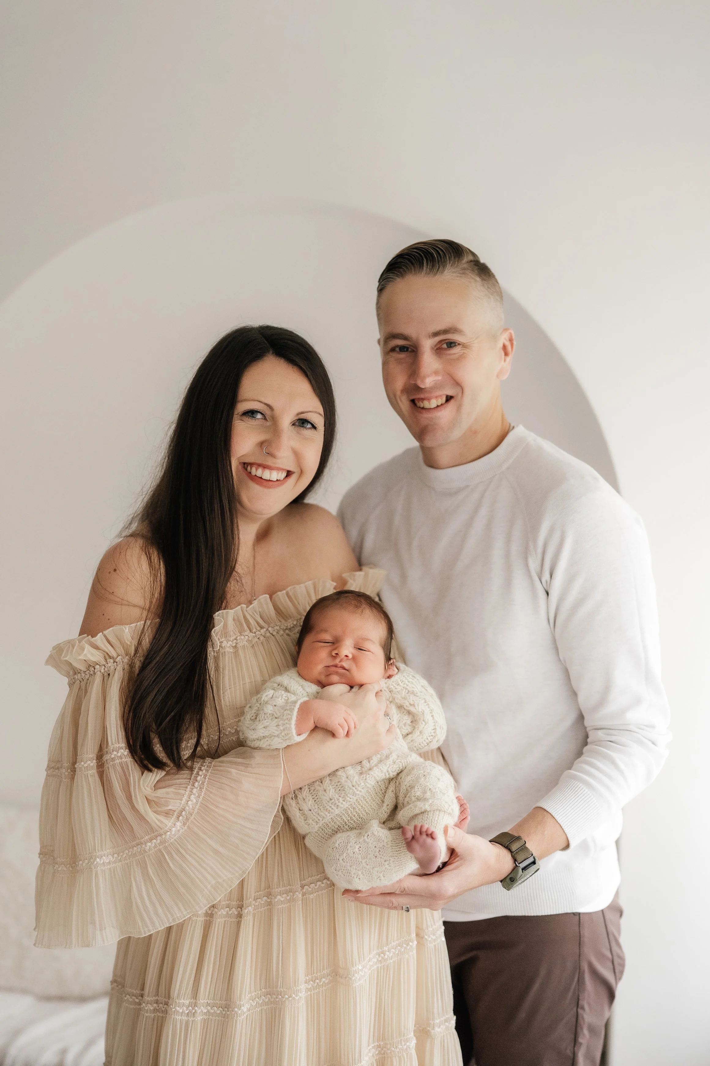 A smiling woman with long dark hair holding a newborn baby dressed in cream-colored knitwear, standing next to a smiling man with short blonde hair, all against a plain light background.