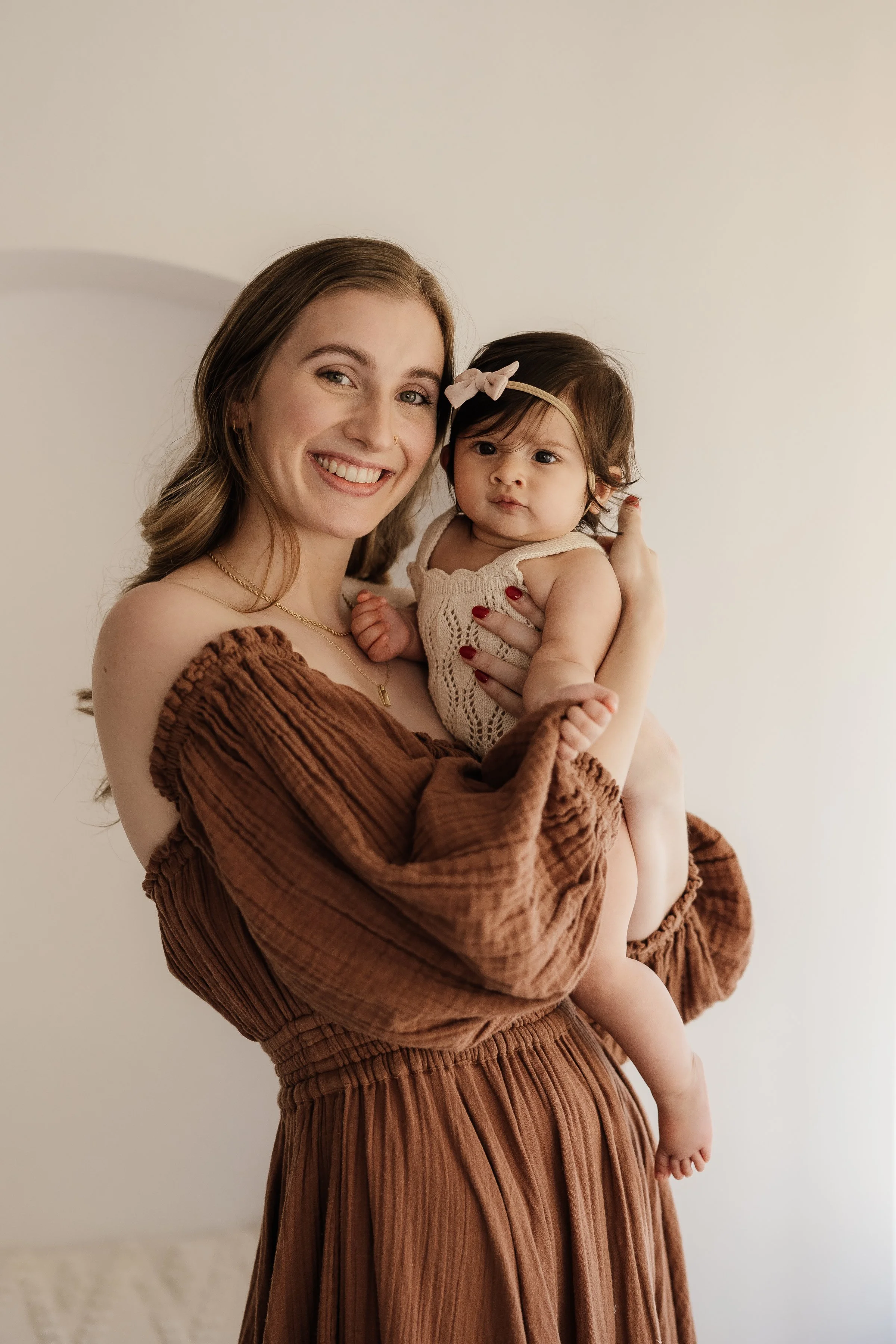 A young woman with long, wavy brown hair and a bright smile is holding a toddler girl. The woman is wearing an off-shoulder brown dress with puffed sleeves and the girl is dressed in a cream-colored knit top with a pink bow headband. They are standin