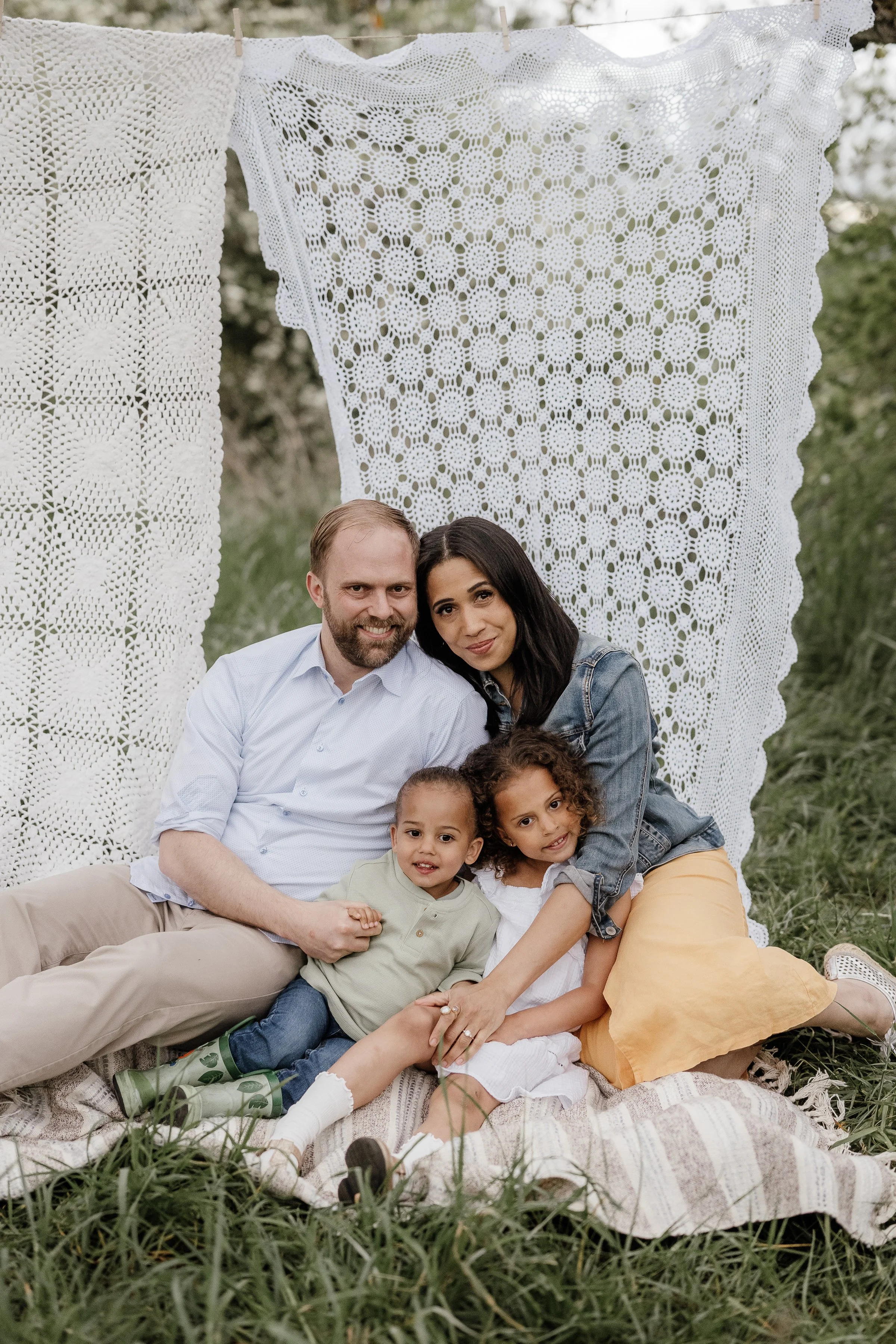 A family of four sitting on a blanket outdoors, with a crochet canopy backdrop, enjoying a day together.