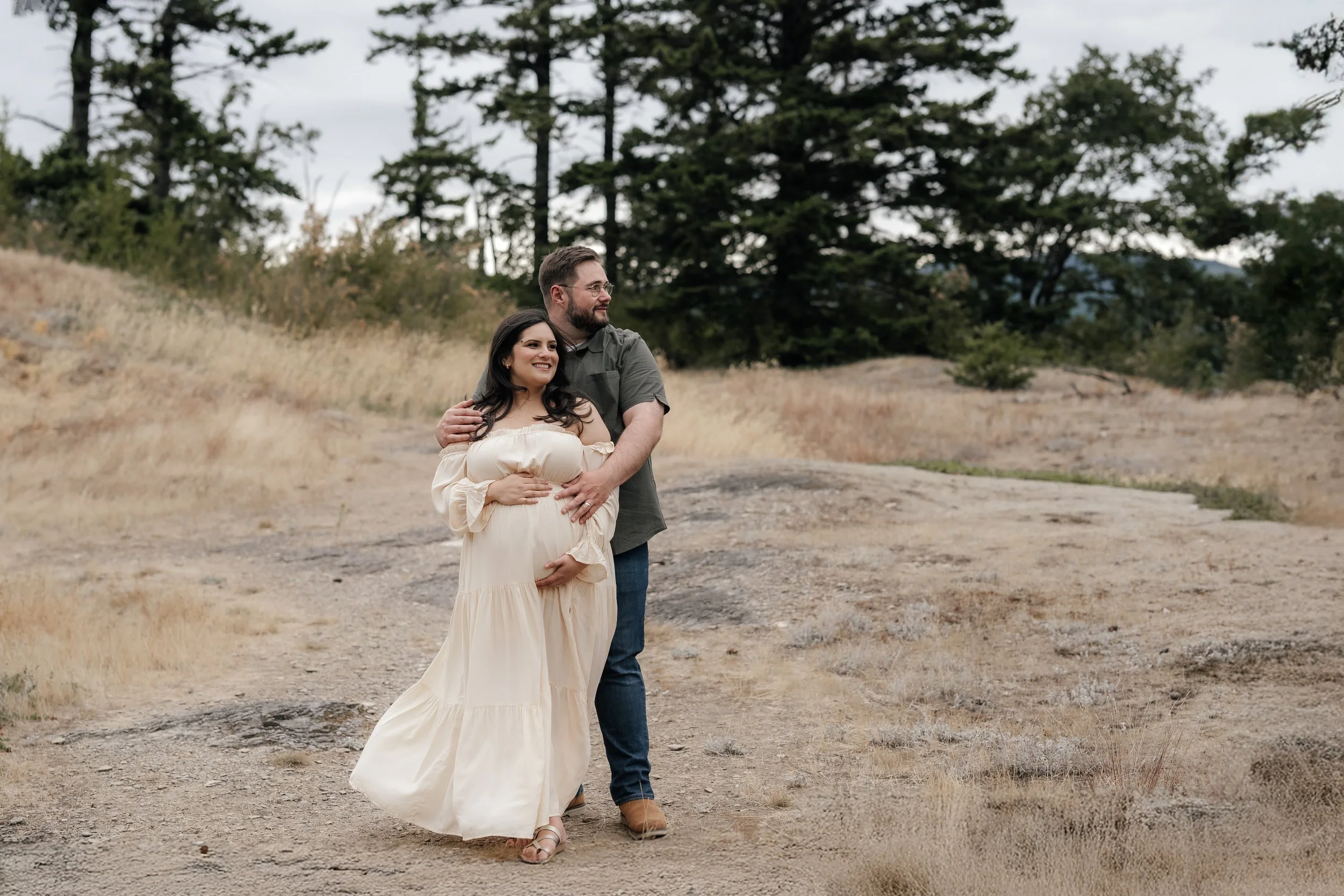 A pregnant woman in a cream-colored dress standing with a man in a green shirt outdoors on a dirt path with trees in the background, smiling and embracing.