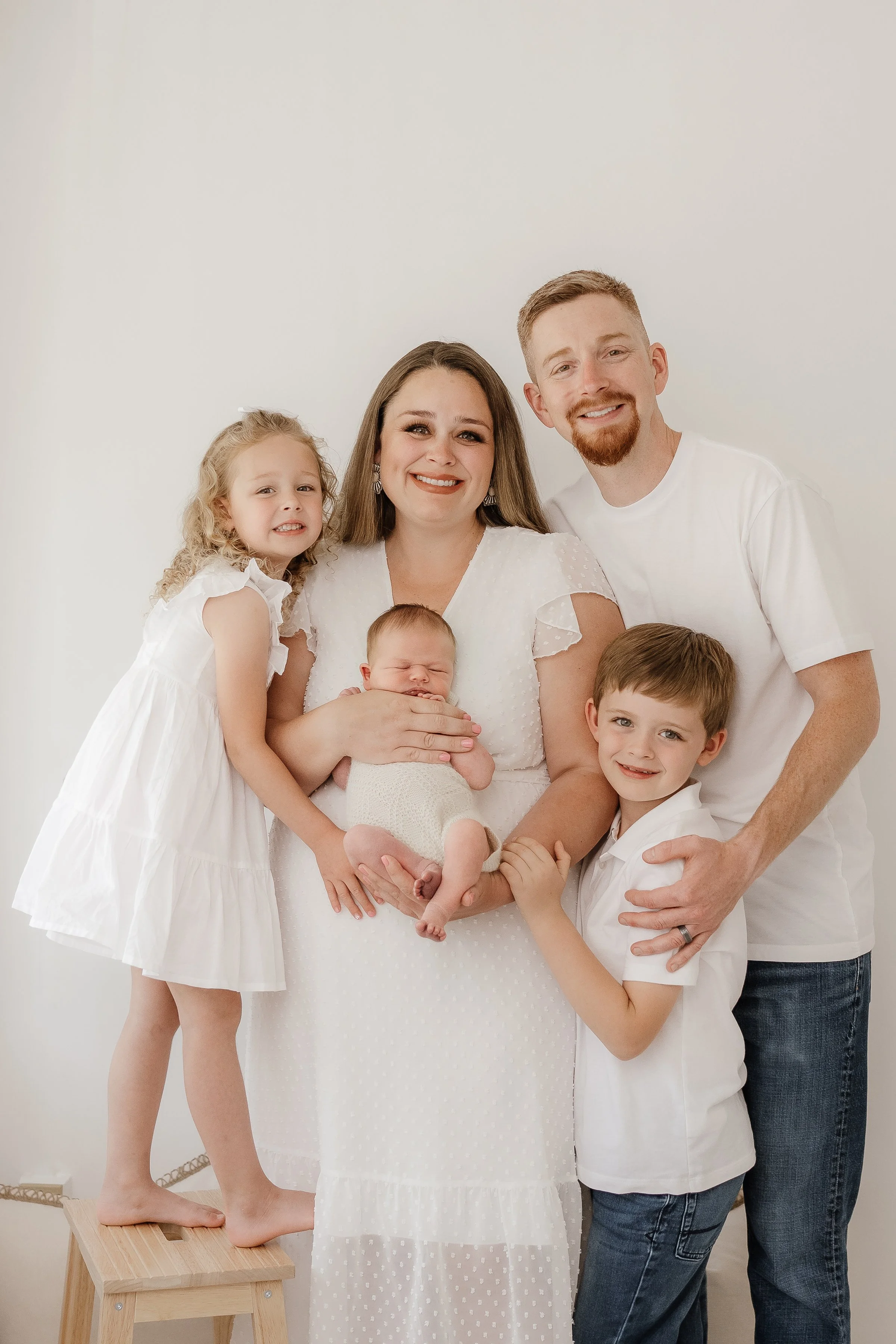 Family of five, including a woman holding a newborn, a young girl standing on a stool, a young boy, and a man, all smiling and dressed in white, posing together against a plain white wall.