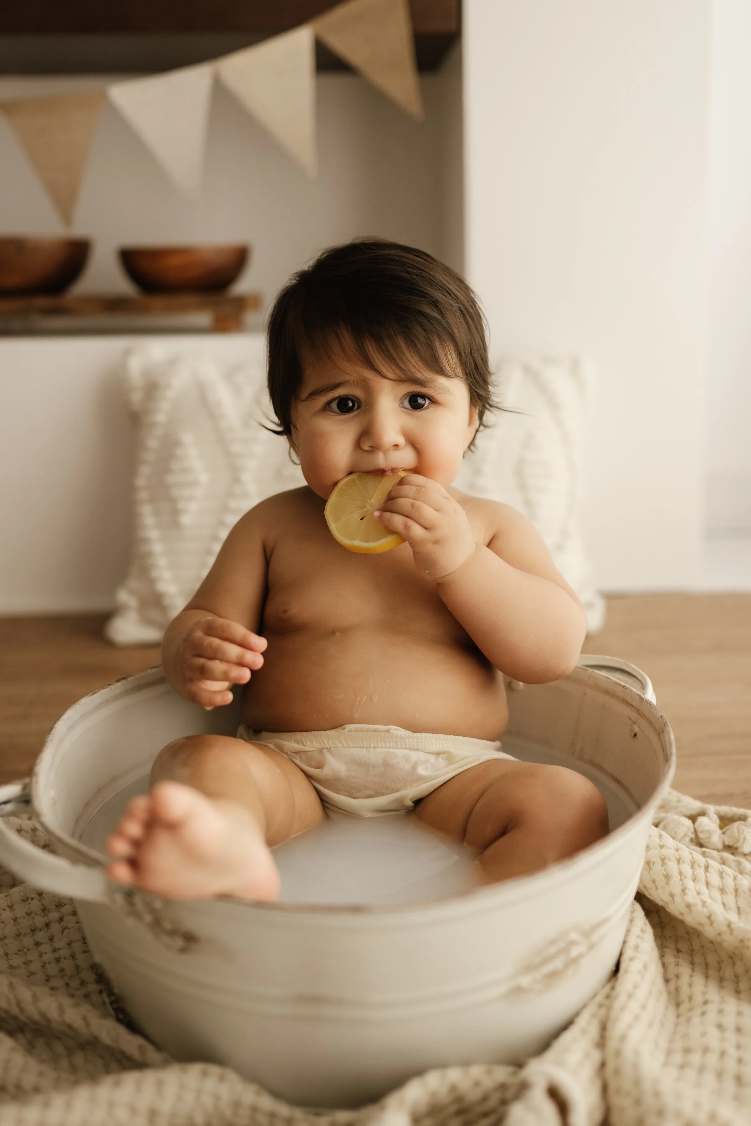 A young child with dark hair, sitting in a white basin filled with water, holding a lemon slice, with a neutral expression, in a cozy room with neutral decor.