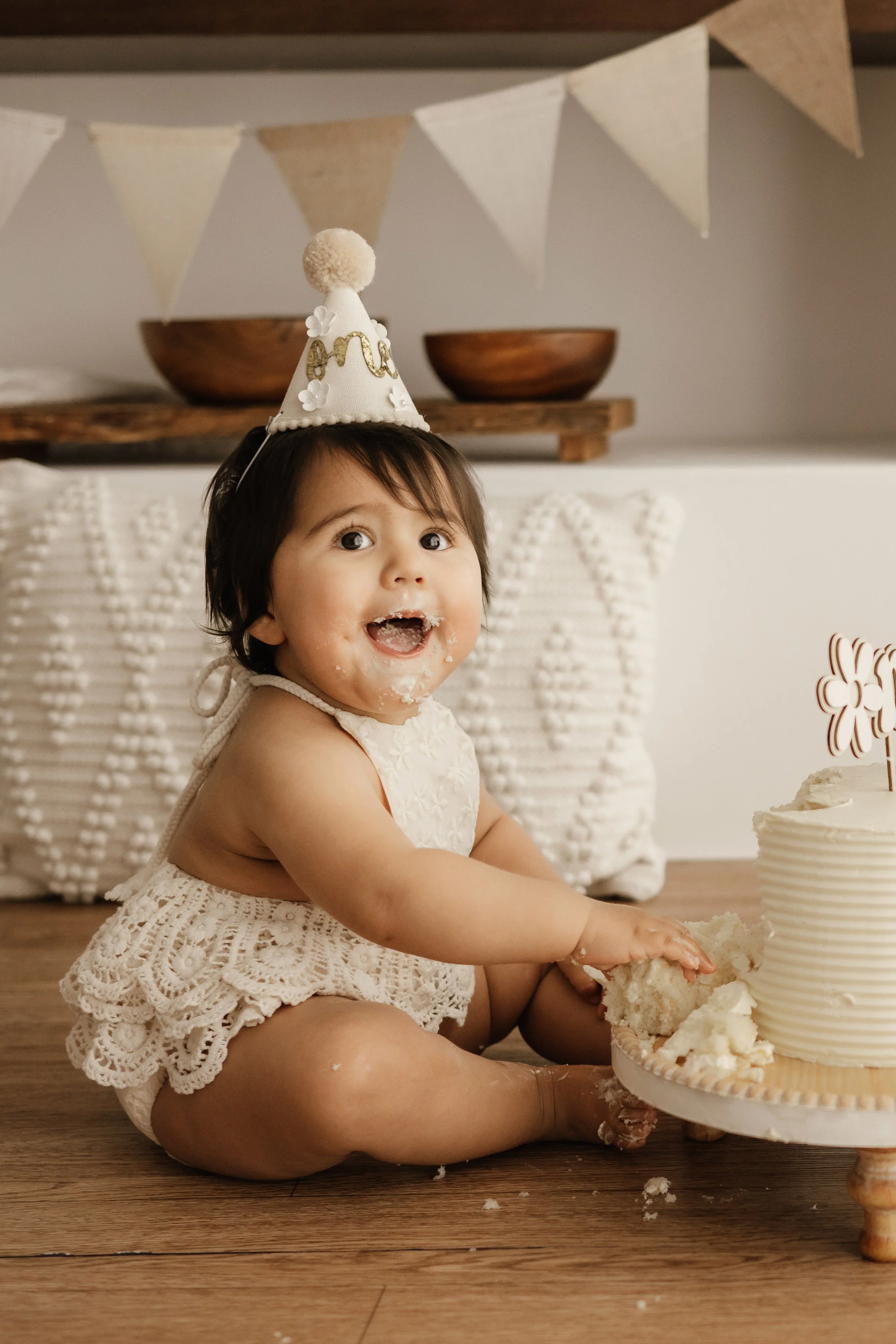 A young girl celebrating a birthday, sitting on a wooden floor, wearing a white lacy dress and a party hat, with cake crumbs on her face and hands, and a partially eaten birthday cake in front of her.