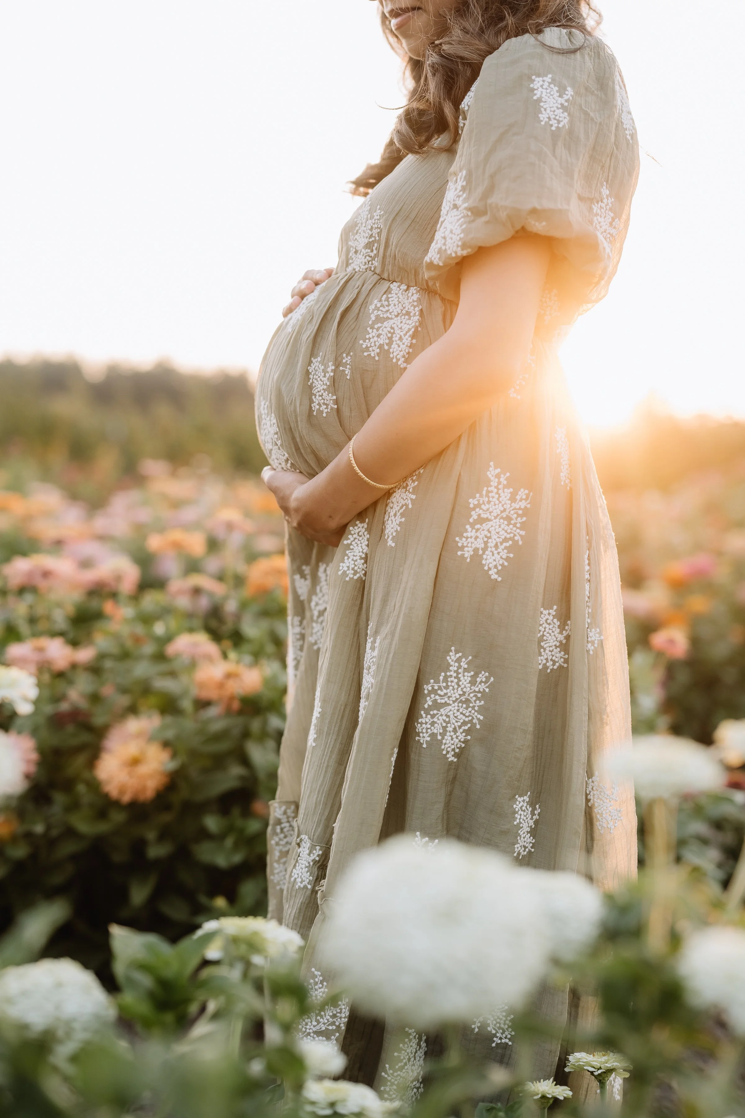 A pregnant woman in a beige dress with white embroidery stands in a field of flowers during sunset, cradling her belly.
