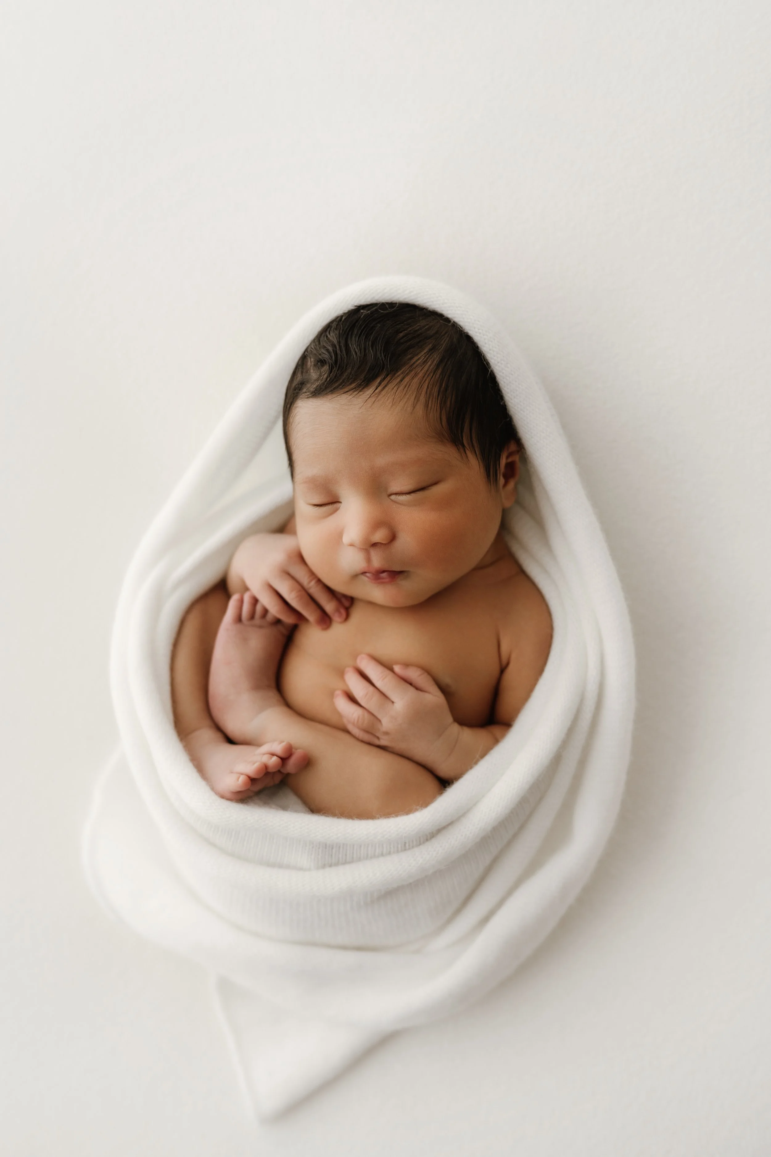 Newborn baby sleeping, wrapped in a white blanket on a white background.