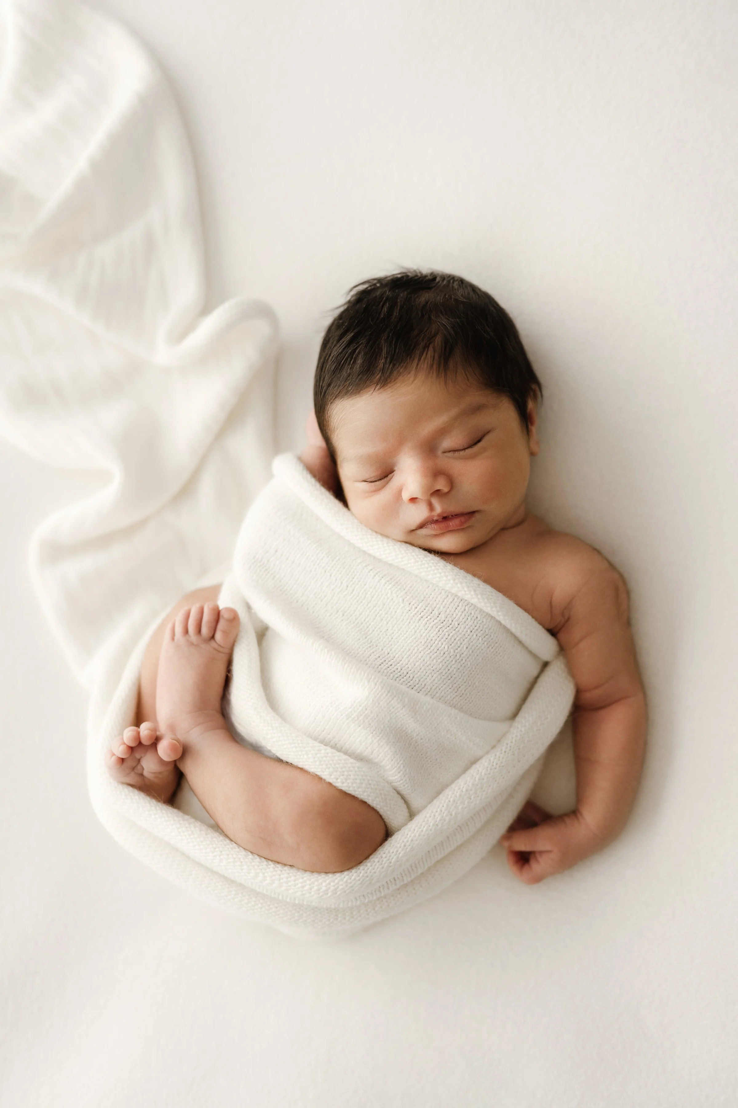 Adorable newborn baby peacefully sleeping, wrapped in a cream blanket, lying on a soft white surface.