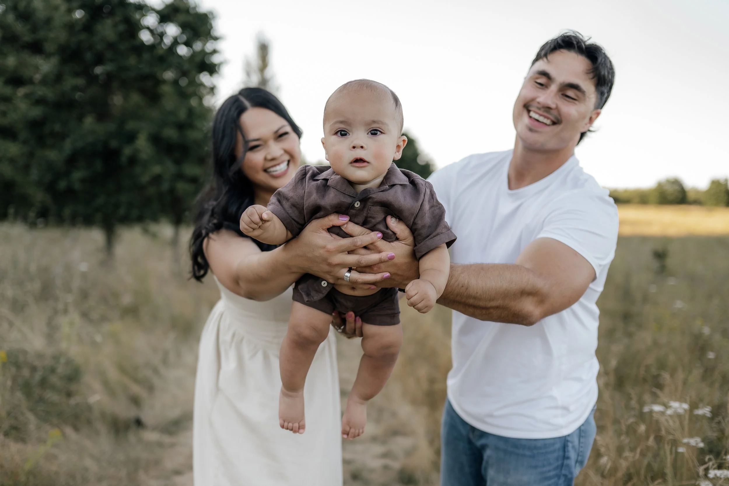 A happy family of three enjoying time outdoors in a field, with the mother and father holding their baby boy between them.