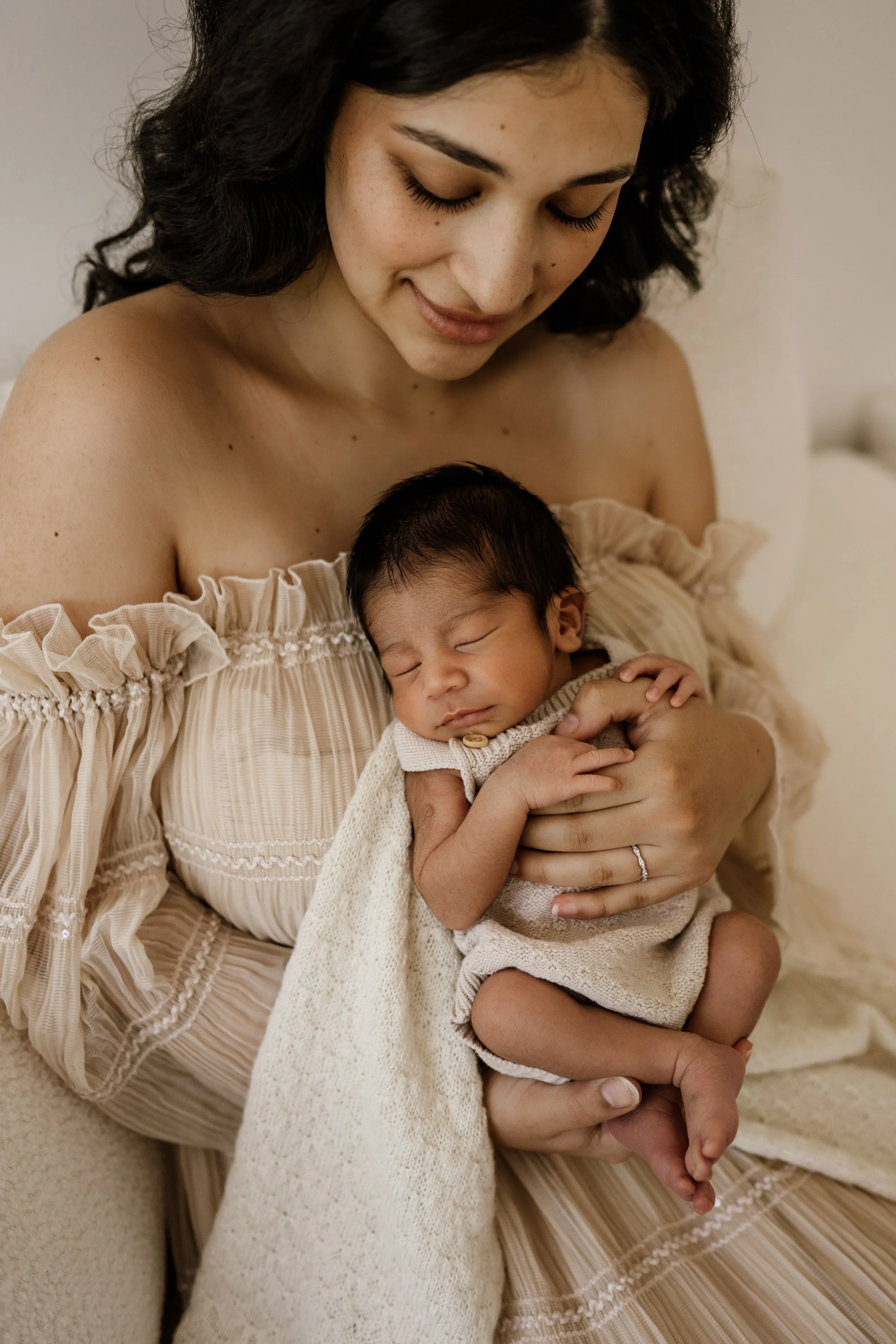 A woman with dark hair and a beige off-shoulder dress holding a sleeping newborn baby in her arms.