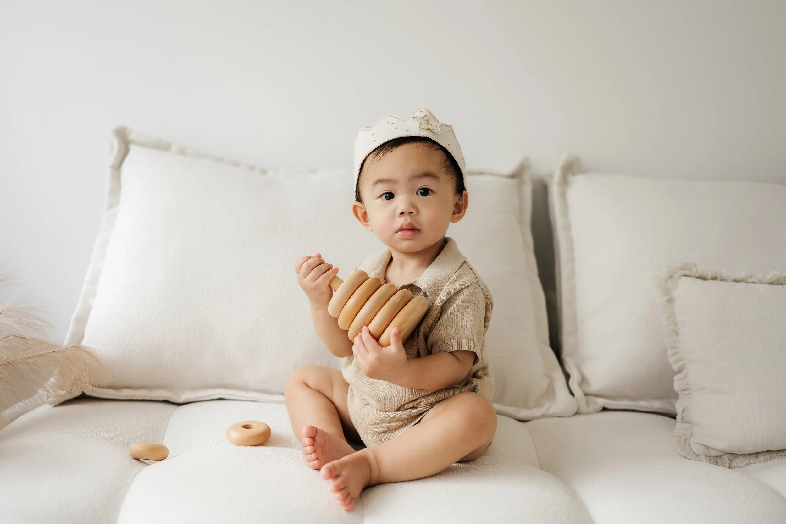 A young Asian boy sitting on a white couch holding wooden stacking rings, wearing a beige outfit and a white hat with a star, with extra rings on the couch.