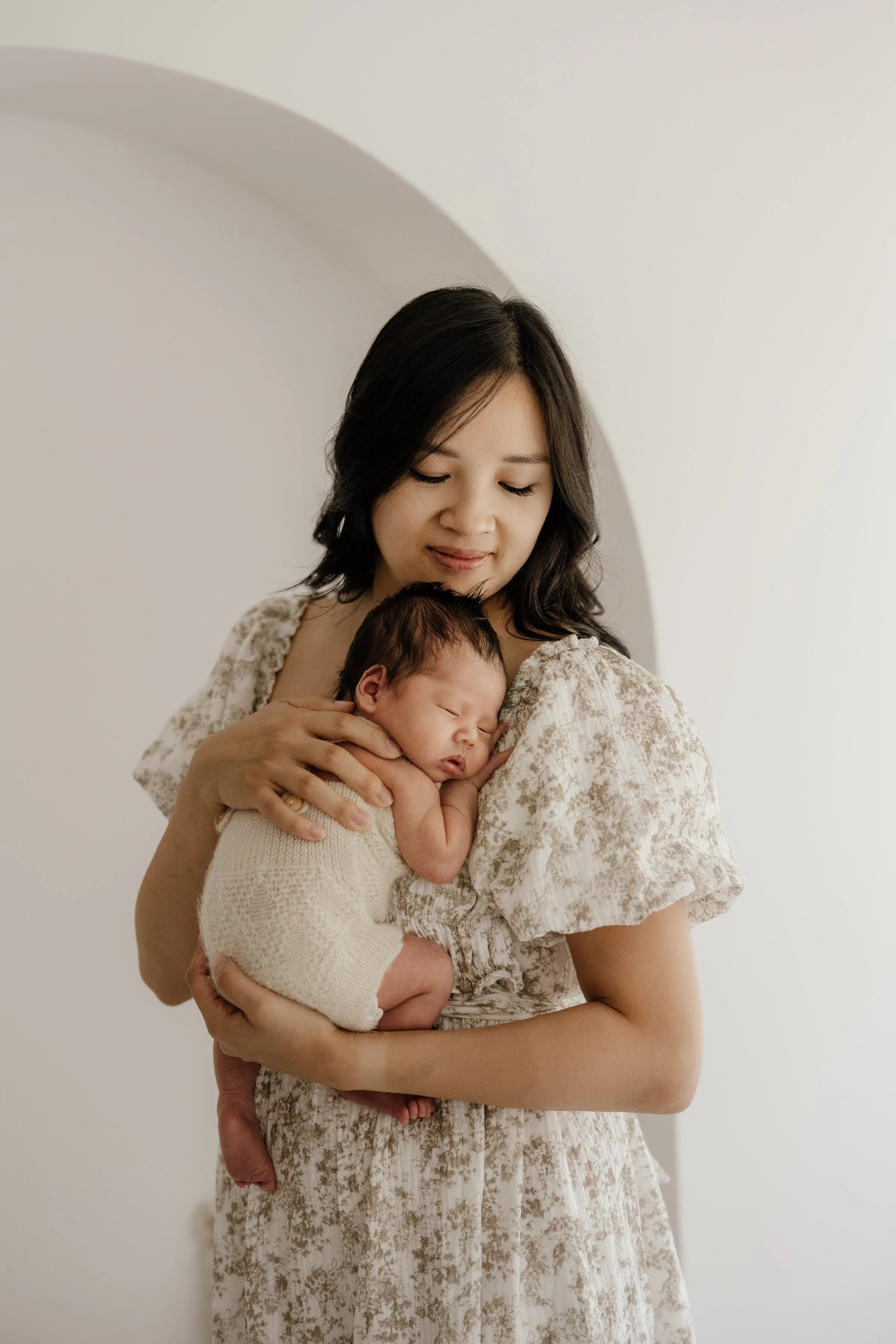A woman gently holding a sleeping newborn baby against her chest, standing in a minimalistic white room.