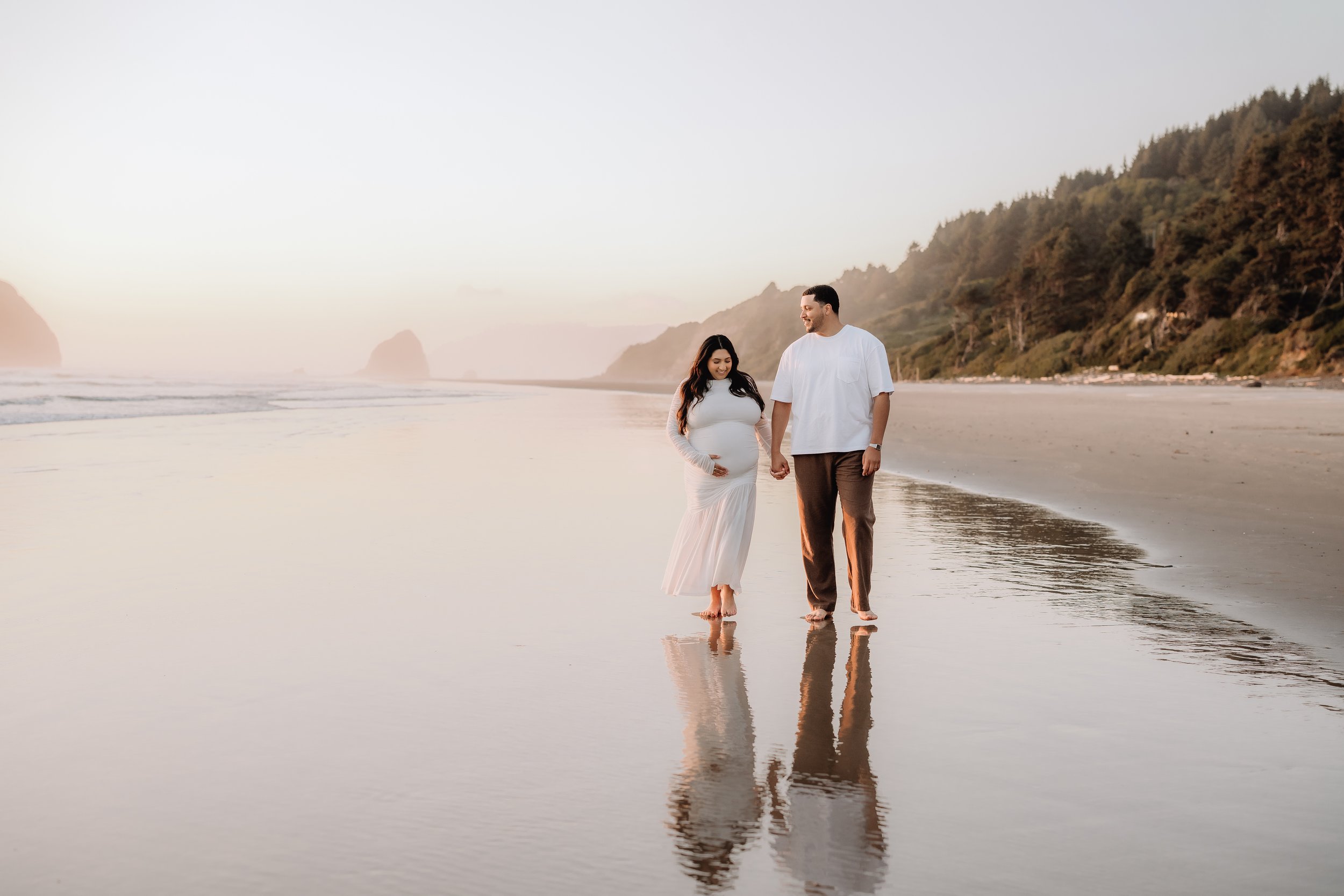 A pregnant woman and a man holding hands walking barefoot on a beach at sunset, with cliffs and hills in the background.