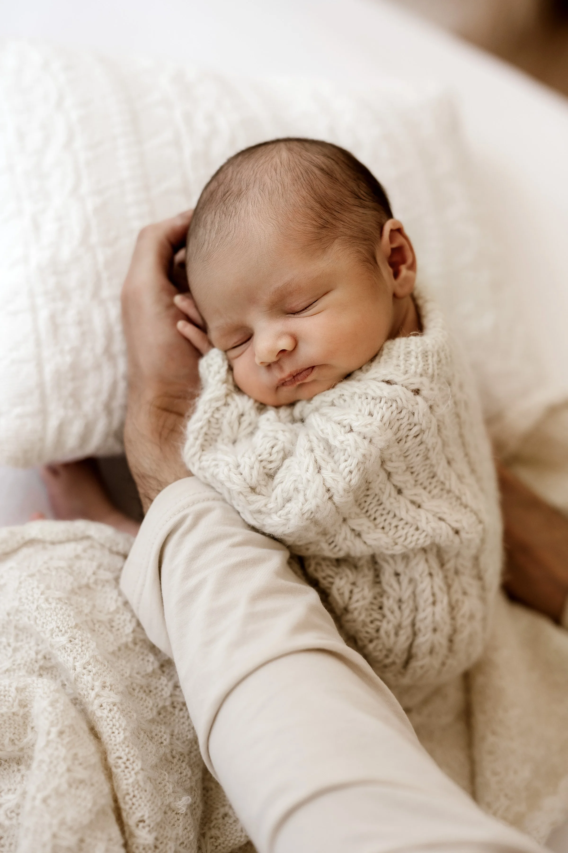 Close-up of a sleeping newborn baby in a cozy cream-colored sweater, resting on a soft blanket, being gently held.