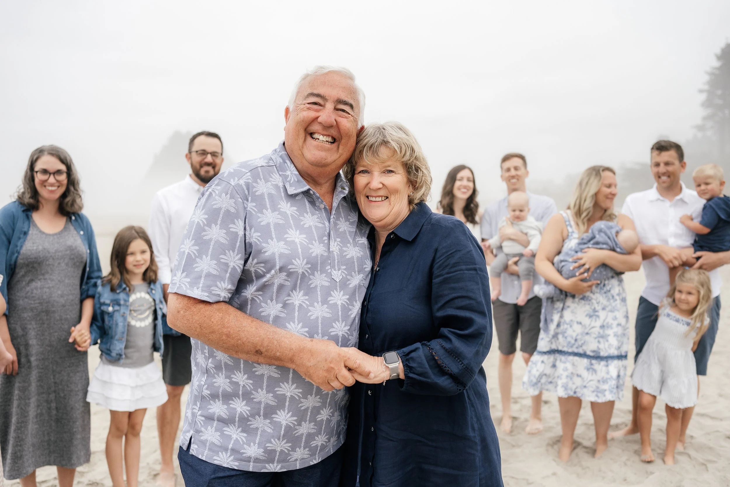 Smiling elderly couple holding hands on a beach with a large family in the background.