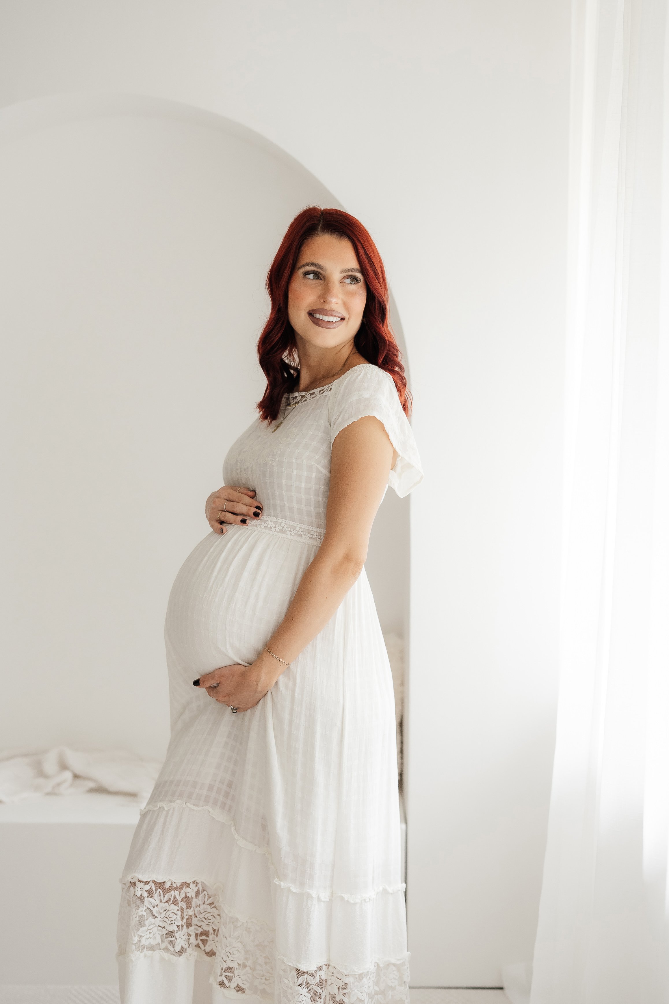 Pregnant woman with red hair wearing a white dress, smiling, and standing in a bright, minimalist room.