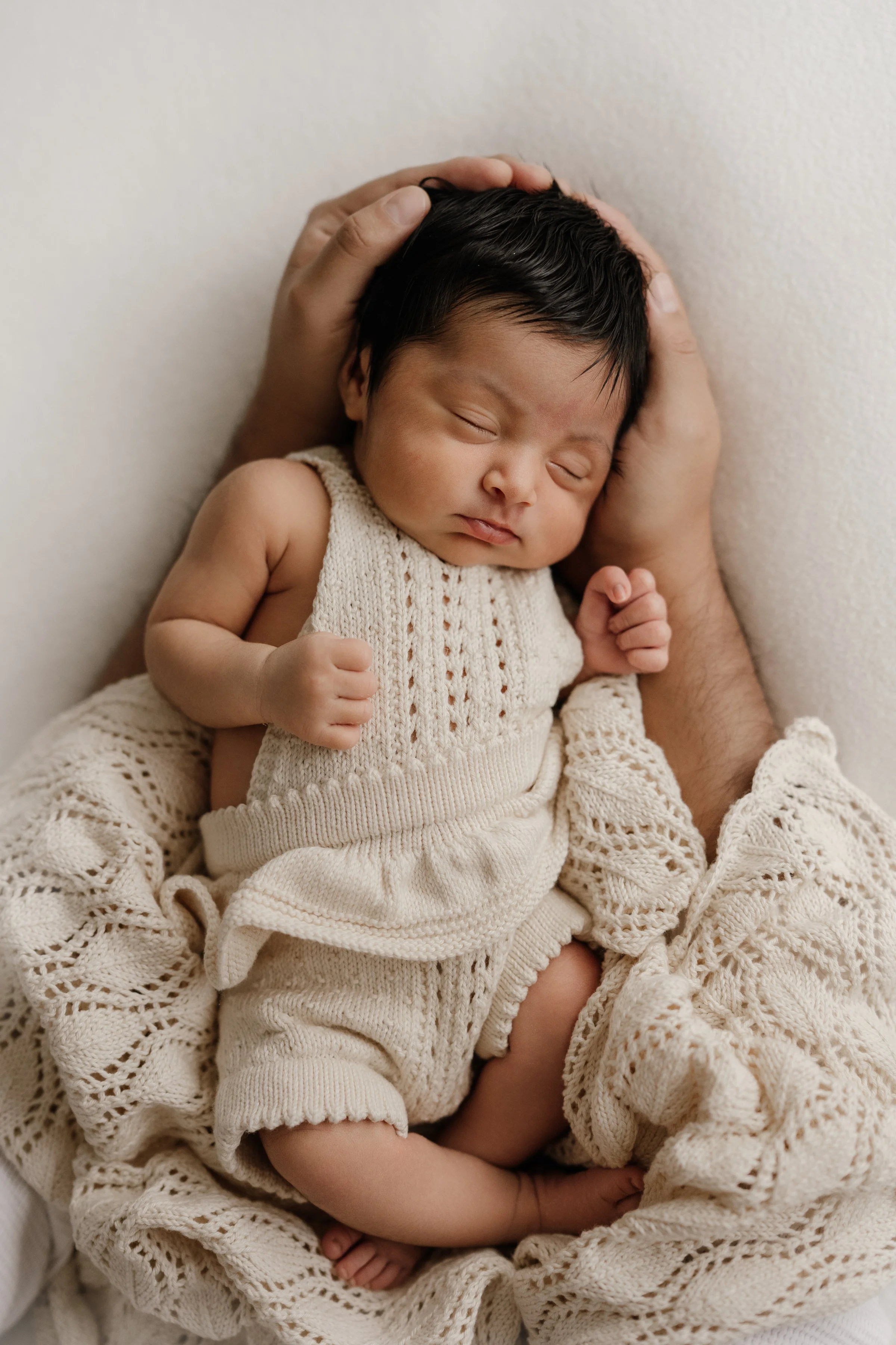 A newborn baby with dark hair sleeping peacefully in an adult's arms against a cream-colored background, dressed in cream knit clothing.
