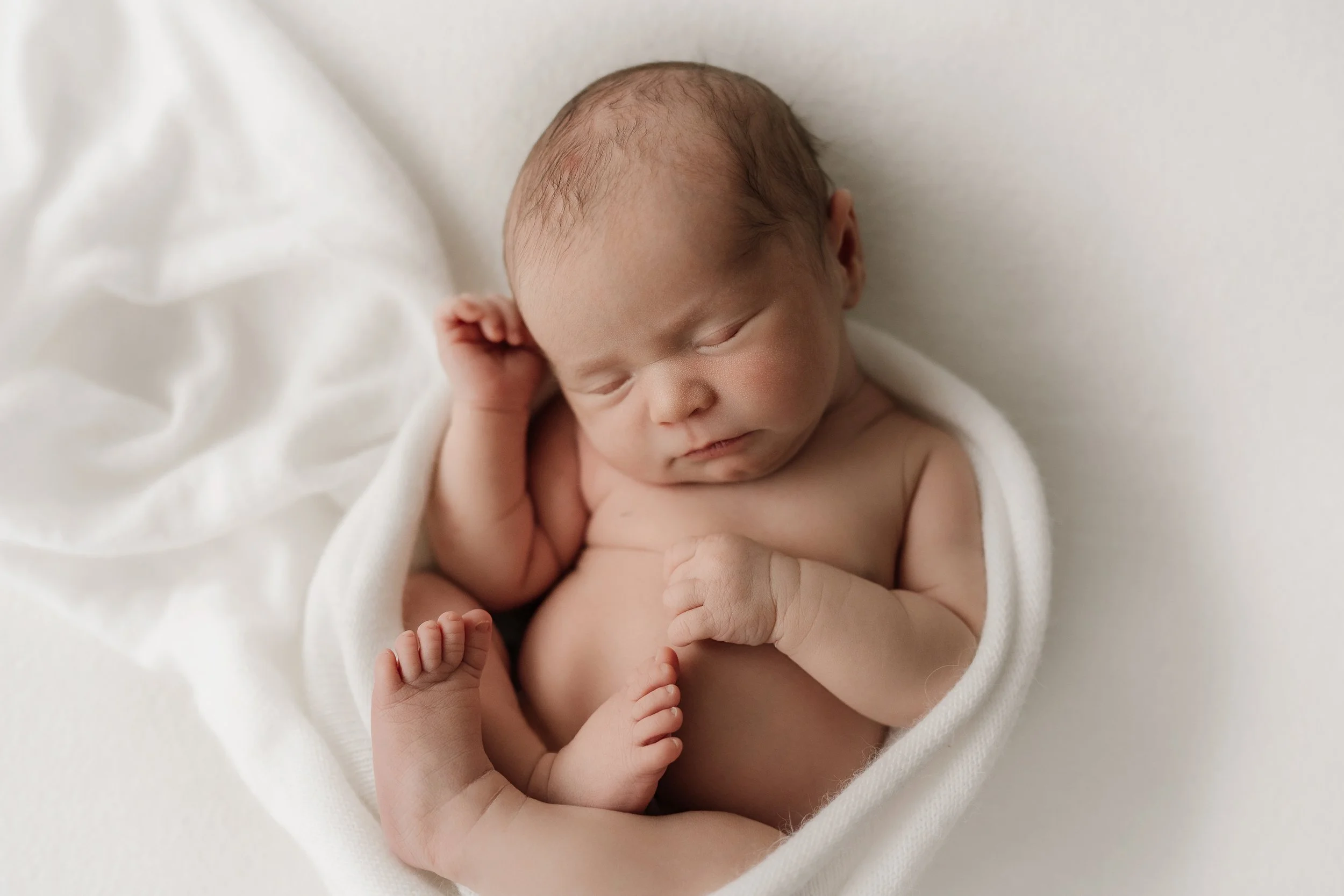 Newborn baby sleeping on a soft white blanket, wrapped partially in a white cloth.