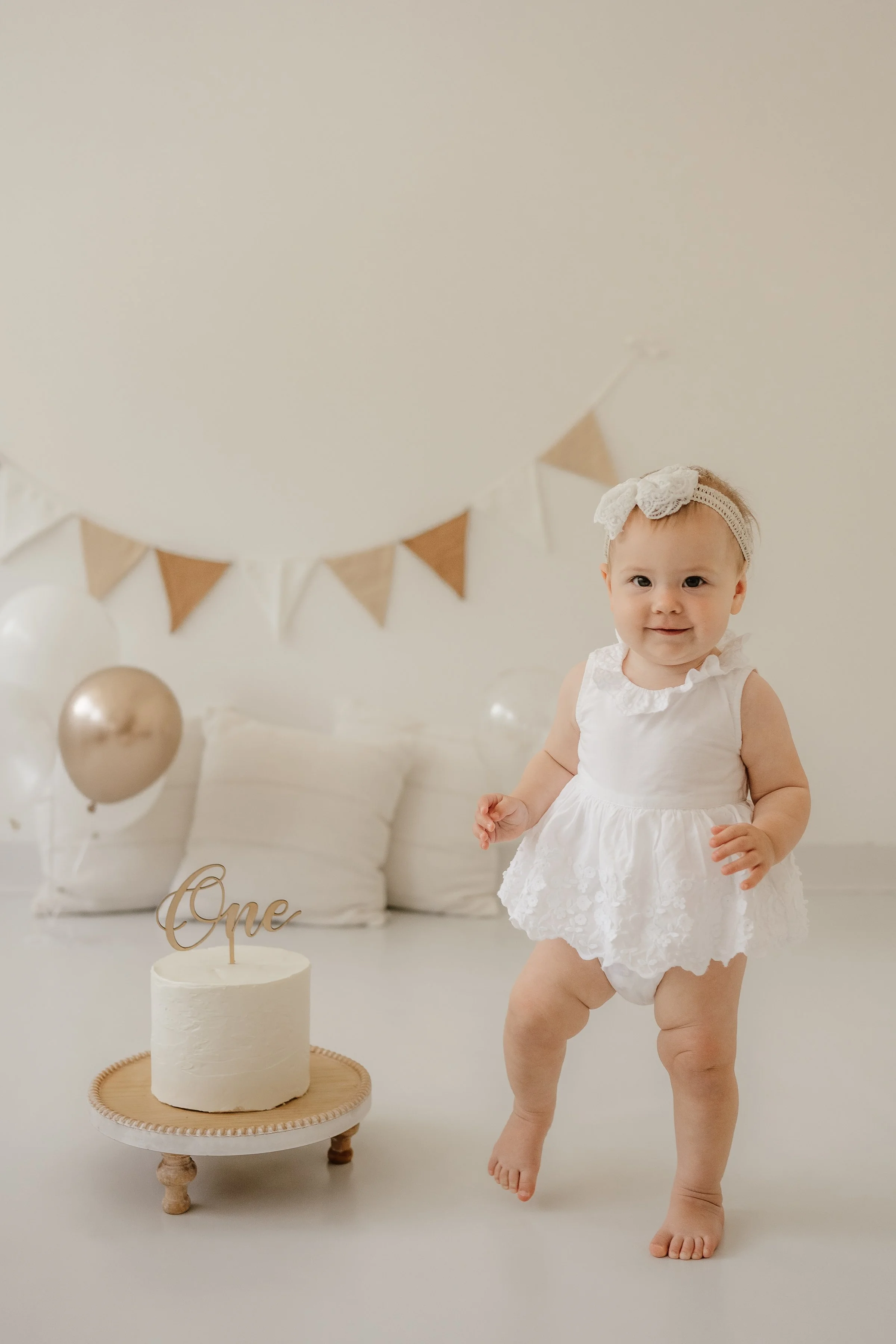 A young girl in a white dress and headband standing next to a small white cake with a "One" topper. The background has beige and white balloons and bunting.