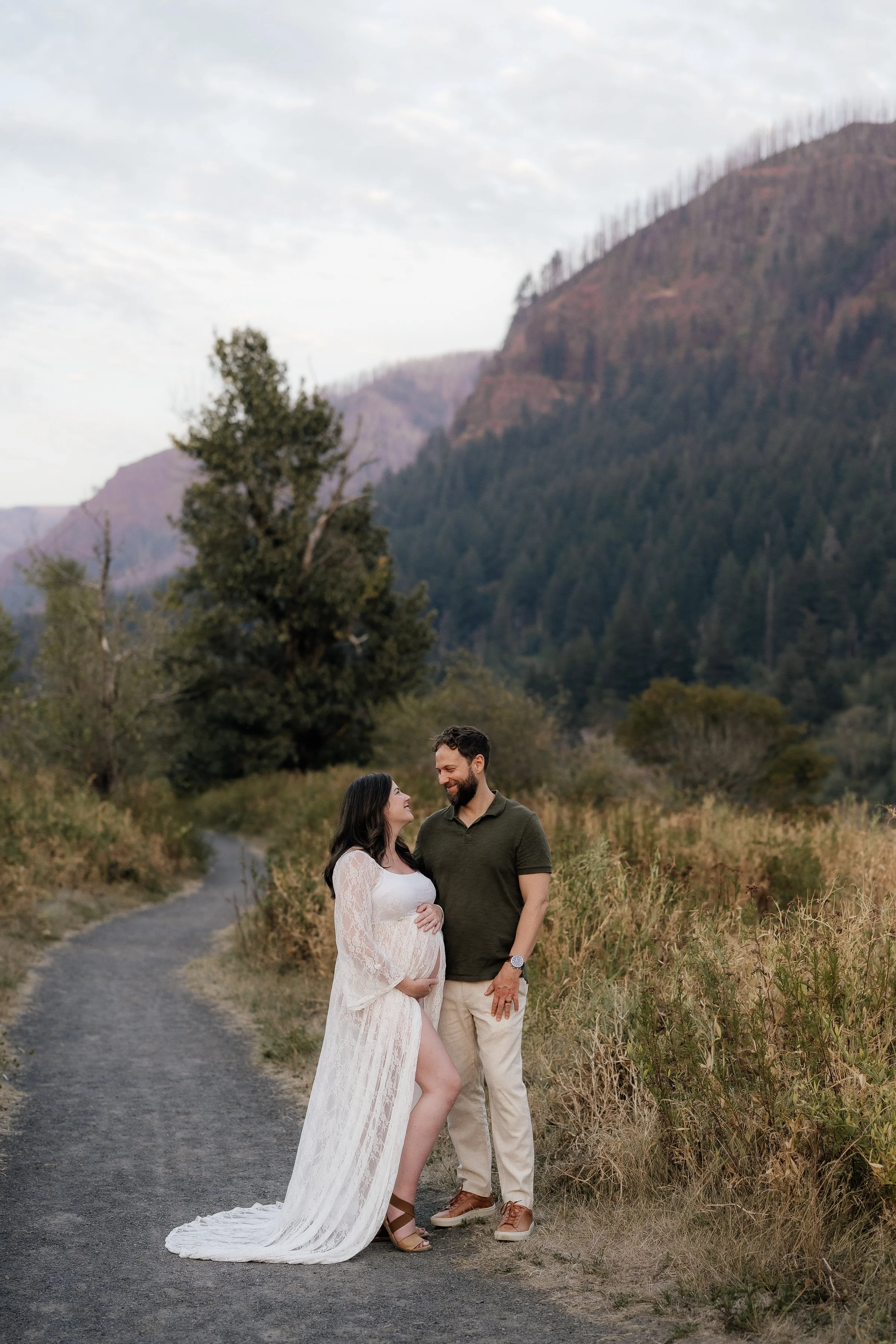 A pregnant woman in a white lace dress stands on a dirt path, smiling at a man in a green polo shirt and beige pants, with a mountain landscape in the background.
