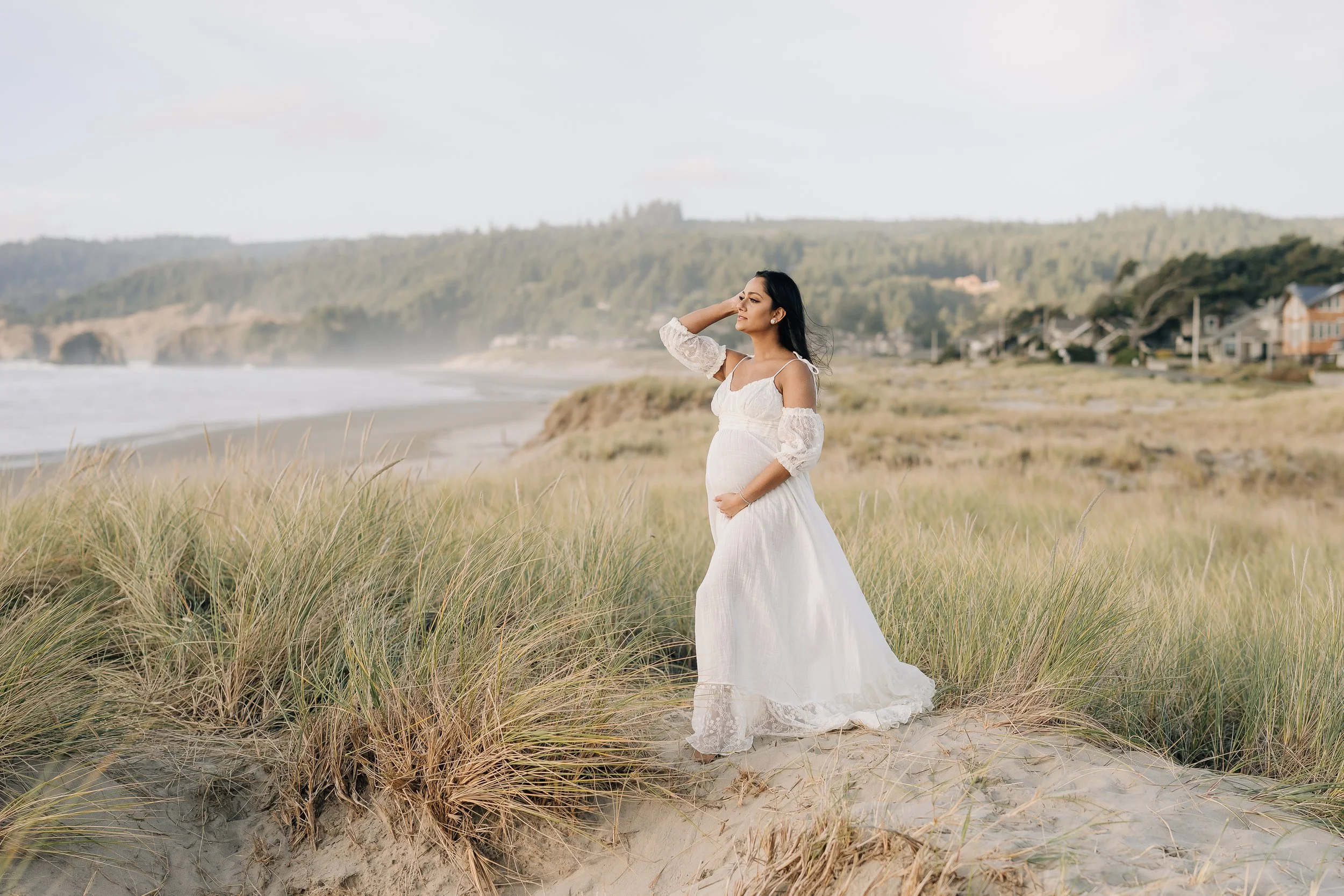Pregnant woman in a white dress standing on a sandy beach with grass, ocean waves, and distant houses in the background.