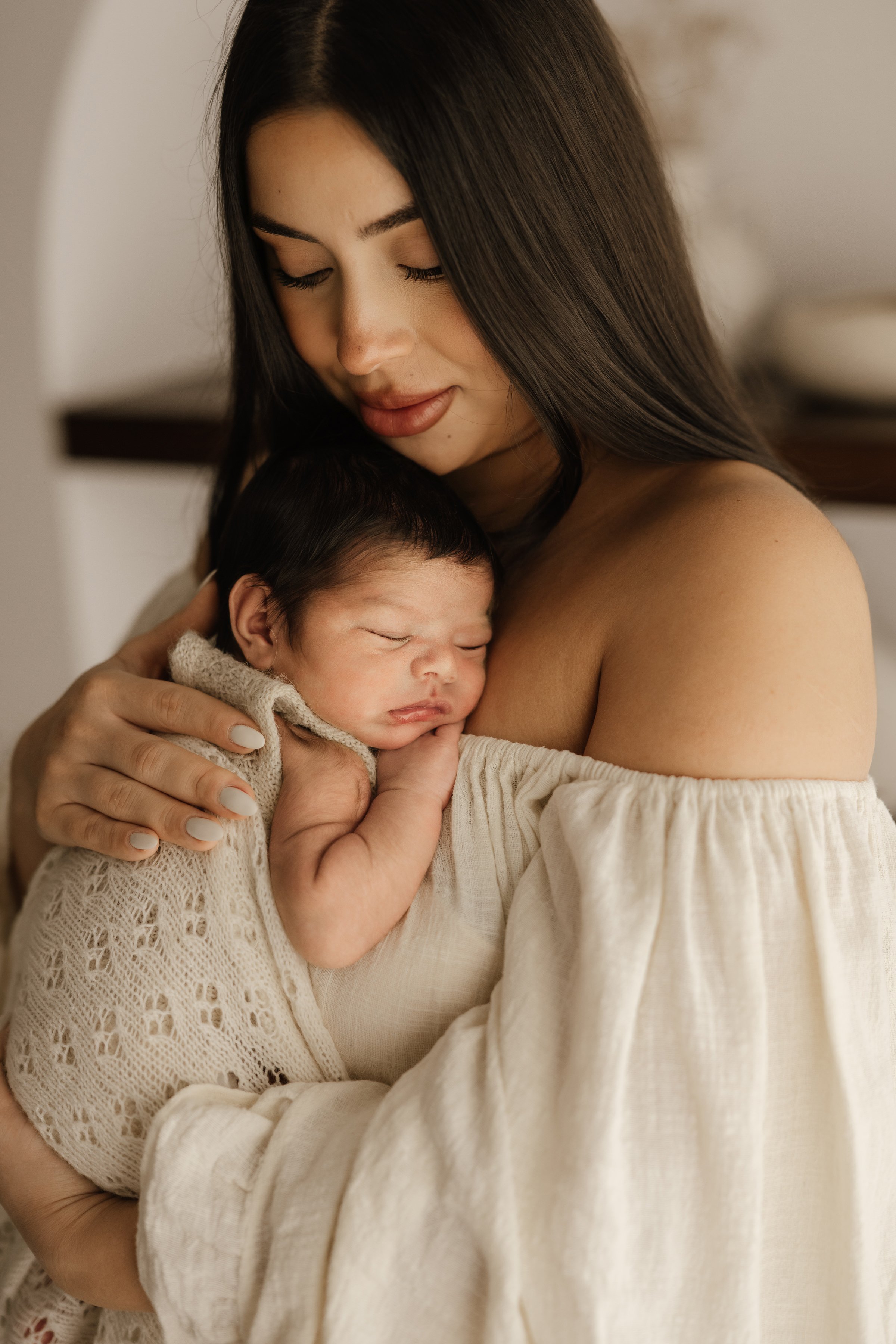 A woman with long dark hair holding a sleeping baby with dark hair against her chest, close to her face.