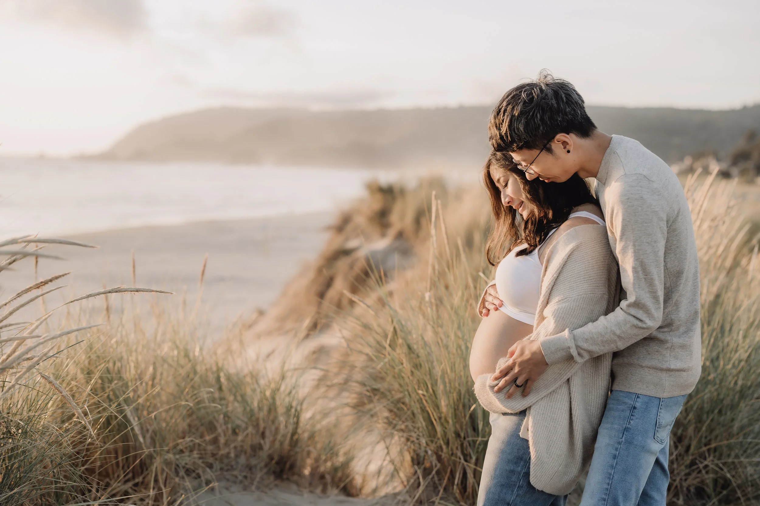 A pregnant woman and her partner embrace on a beach with tall grass, ocean, and distant hills in the background.