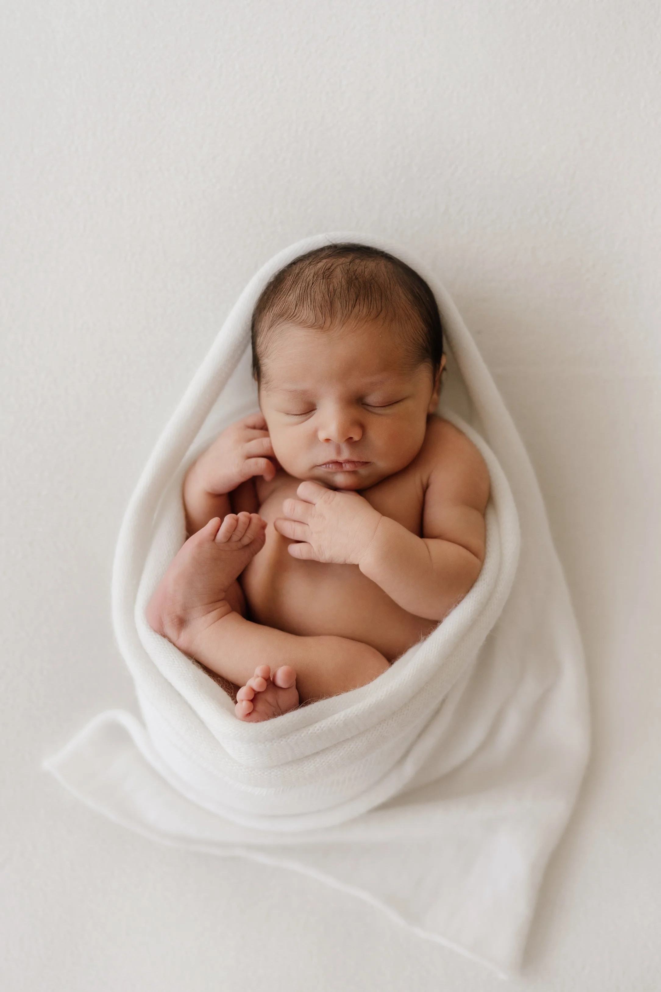 Newborn baby sleeping peacefully, wrapped in a white blanket, on a white surface.