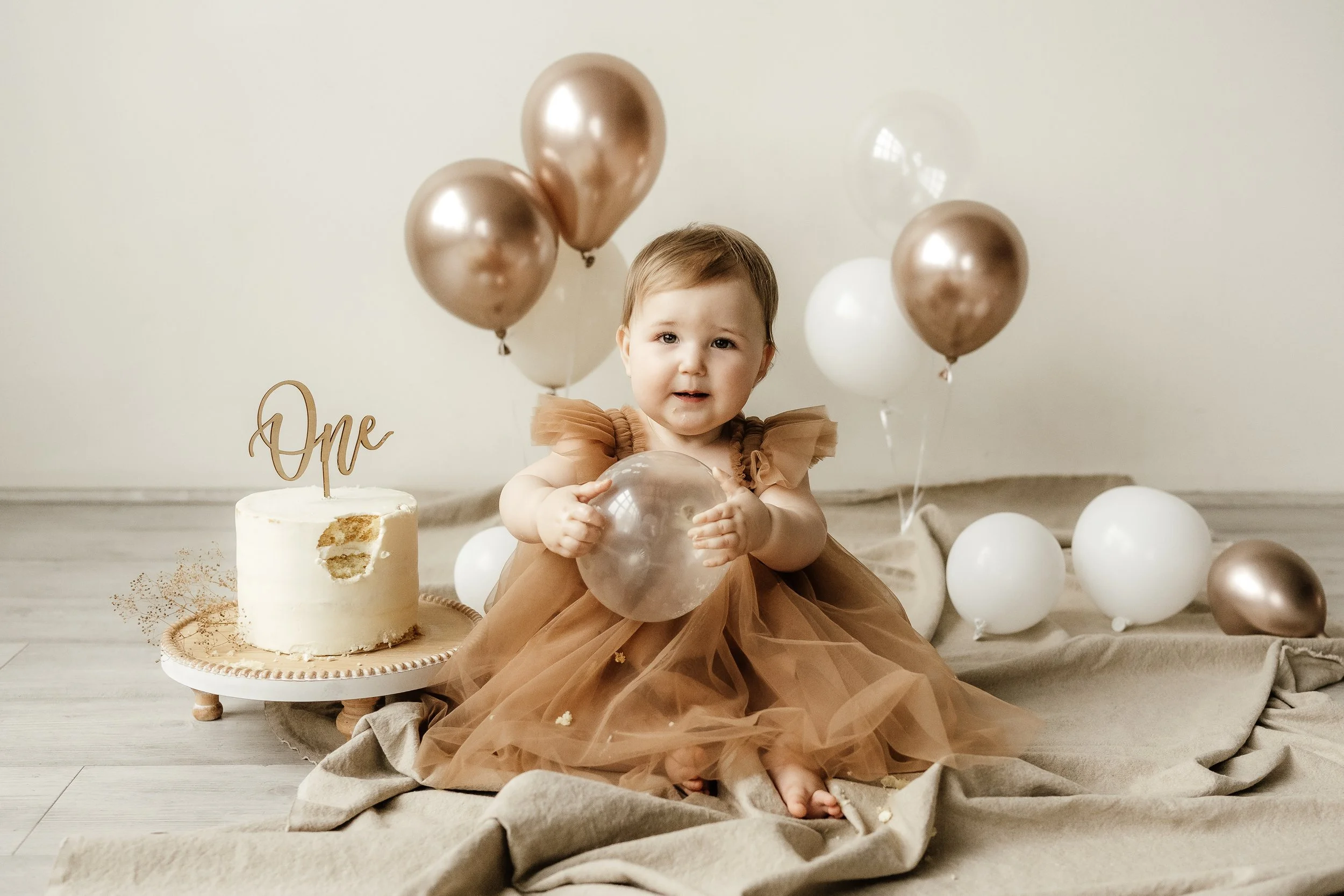A young girl in a brown tulle dress sitting on the floor surrounded by white and bronze balloons, holding a balloon, with a small white cake with a 'One' cake topper behind her, in a neutral-toned setting for her first birthday celebration.