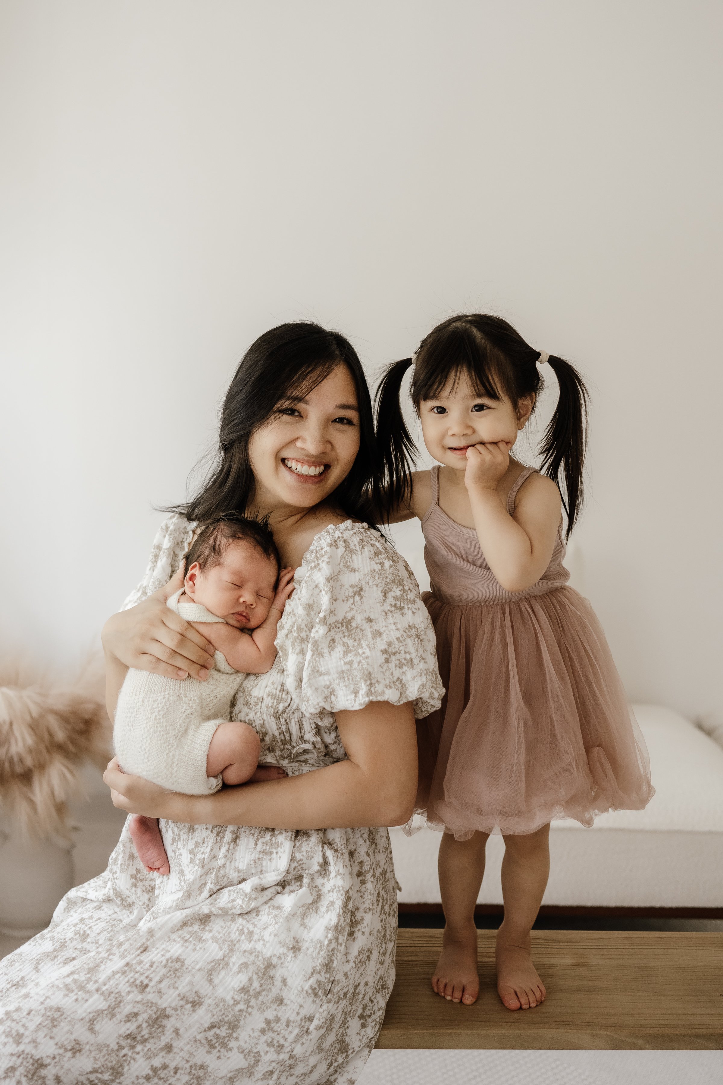 Mother holding a newborn baby while the father stands in the background with their older daughter during an in-home lifestyle newborn session in Portland.