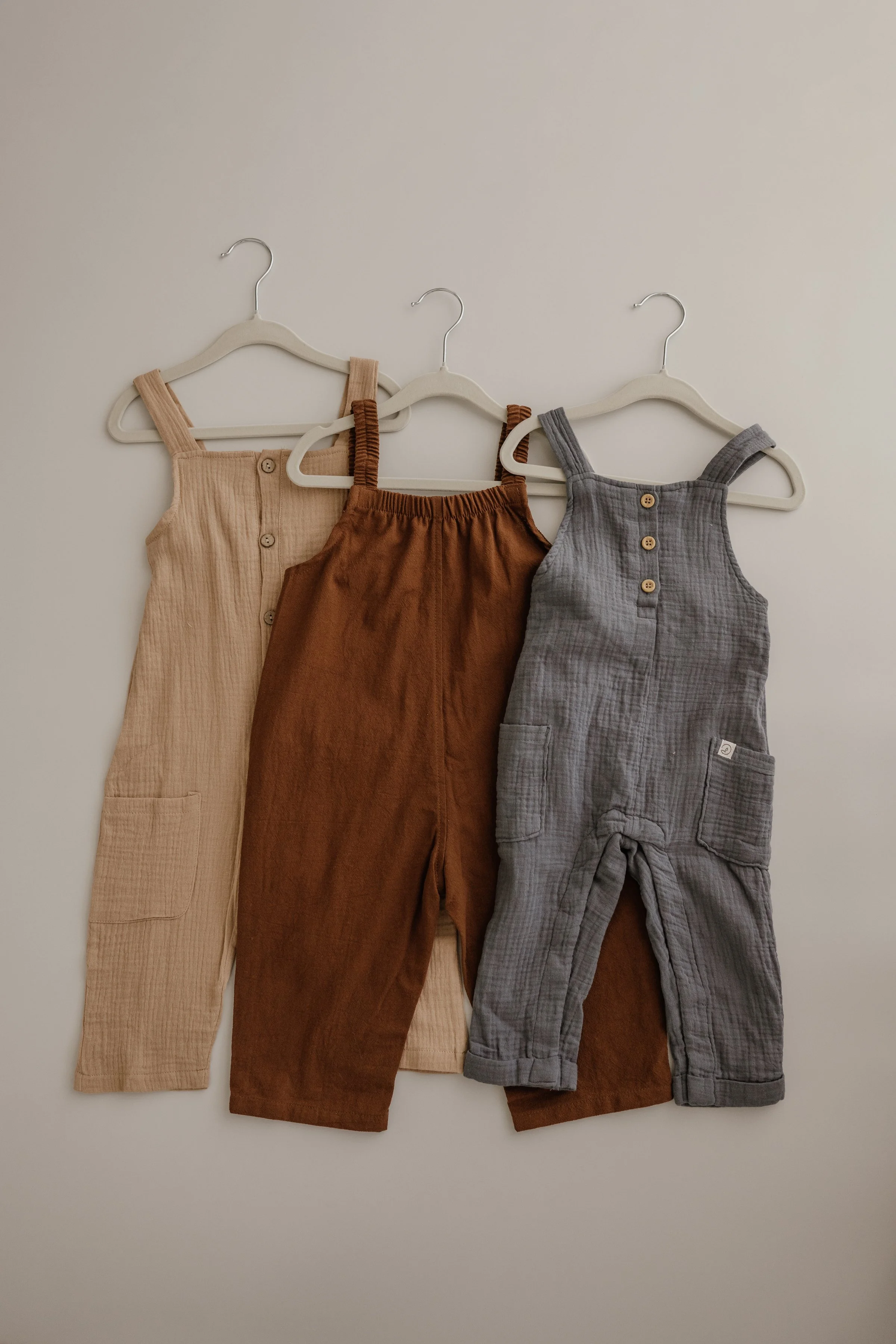 Three children's outfits hanging on hooks against a plain wall, including a beige romper, a brown pair of overalls, and a gray pair of overalls.
