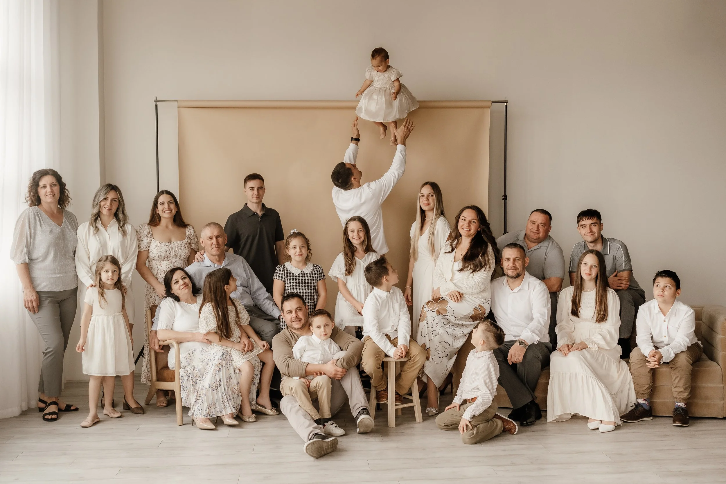 A large multigenerational family photo with children, teenagers, and adults, gathered together in a bright room, with a man lifting a young girl in a white dress in the center.