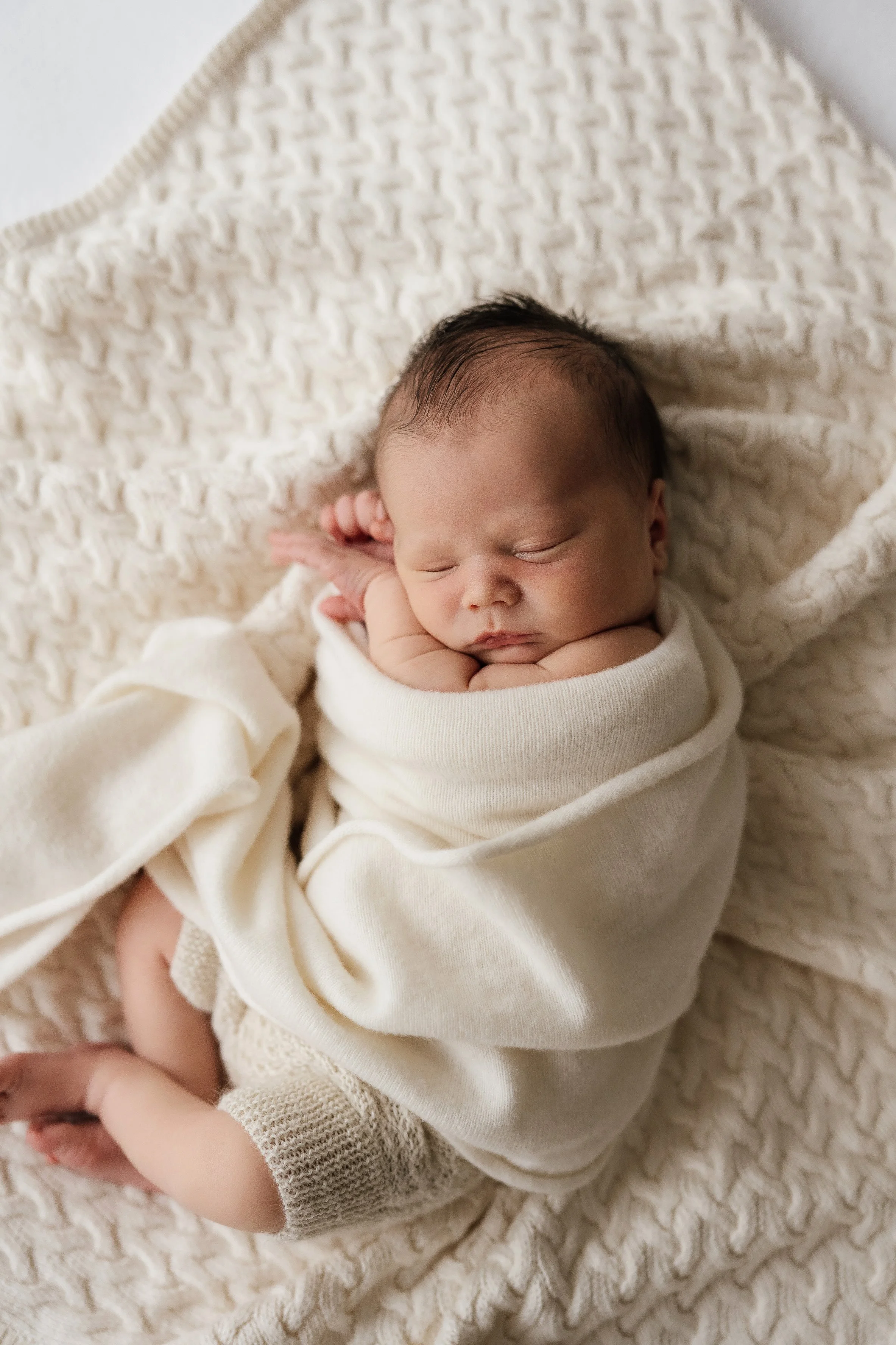 A sleeping newborn baby swaddled in a cream blanket, lying on a textured cream blanket.
