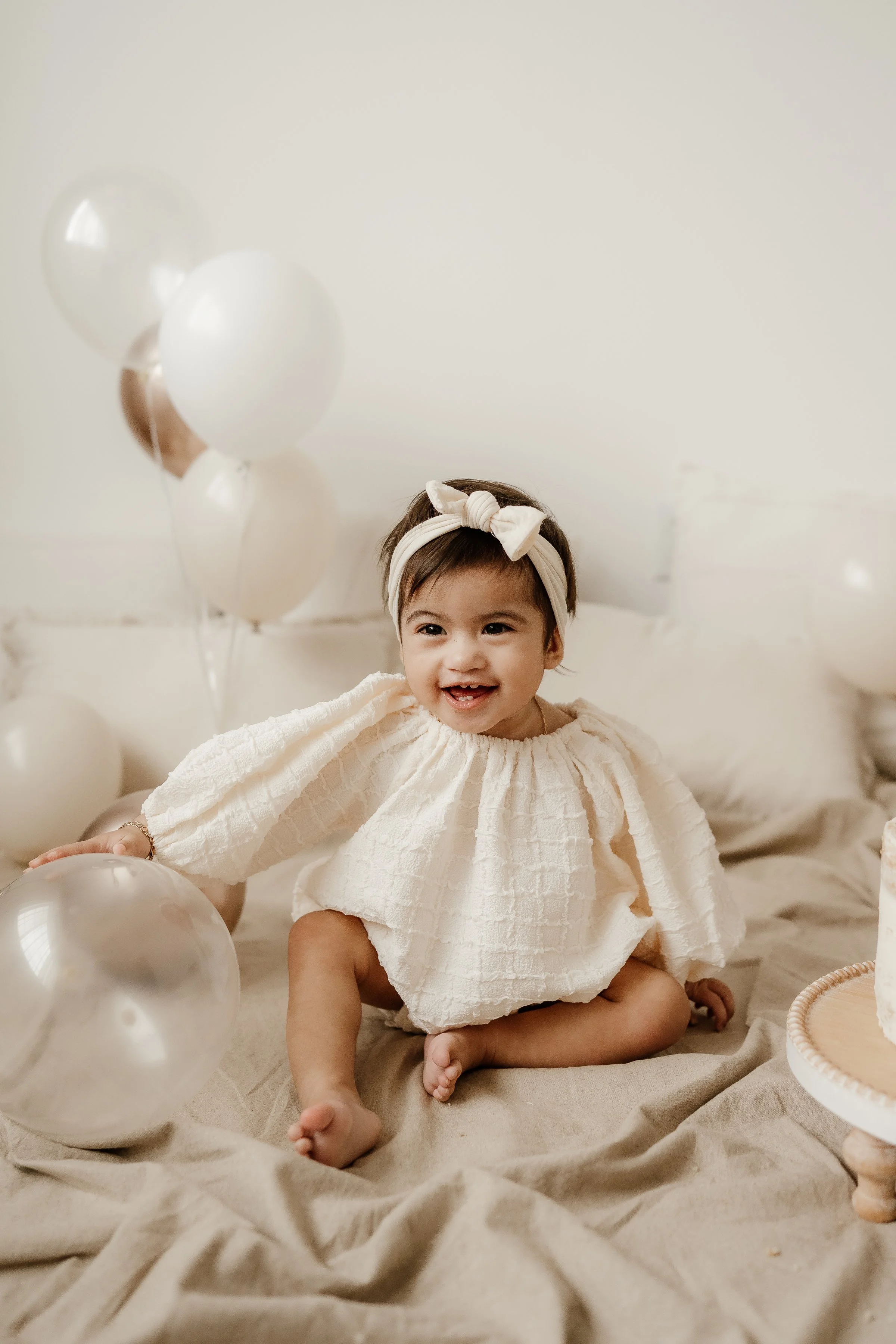 A smiling young girl sitting on a beige blanket, surrounded by white and transparent balloons, wearing a cream-colored textured dress and a white headband with a bow.